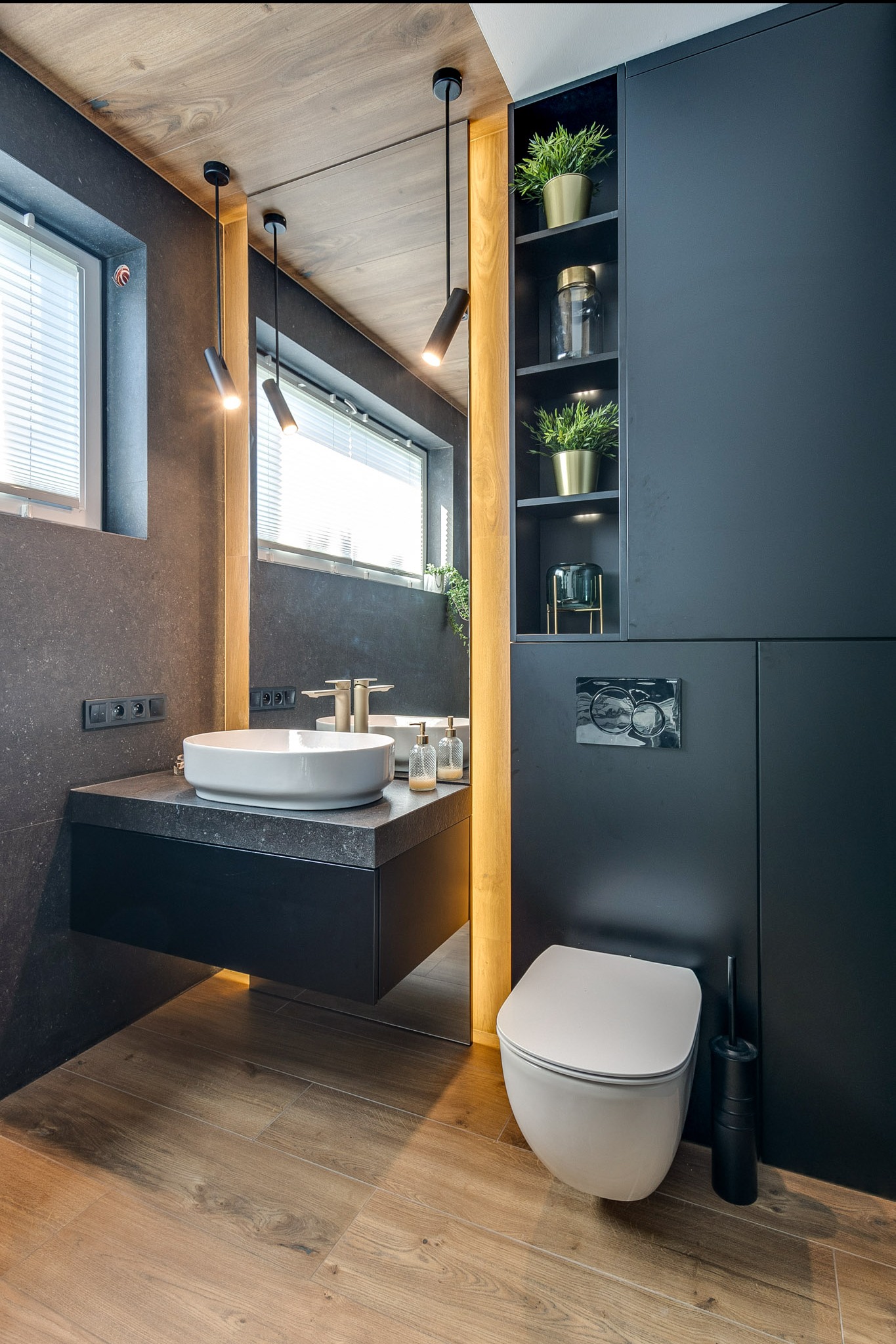 Nowoczesna łazienka z czarnymi szafkami, drewnianym akcentem i białym umywalką. Oświetlenie punktowe i rośliny doniczkowe dodają elegancji. Ujęcie z perspektywy.