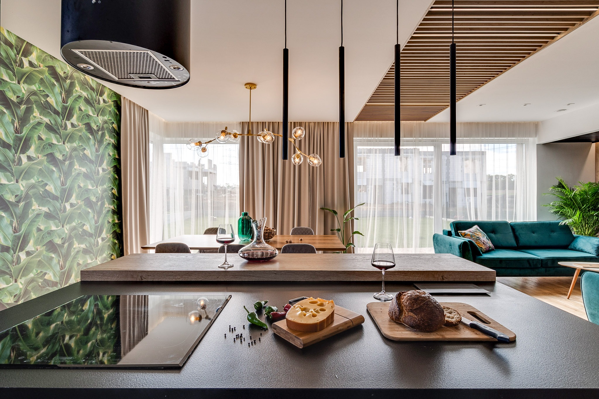 Nowoczesna kuchnia z wyspą, na której leżą ser, chleb i wino. W tle salon z zieloną sofą i tapetą w liście bananowca. Całość doświetlona lampami wiszącymi.