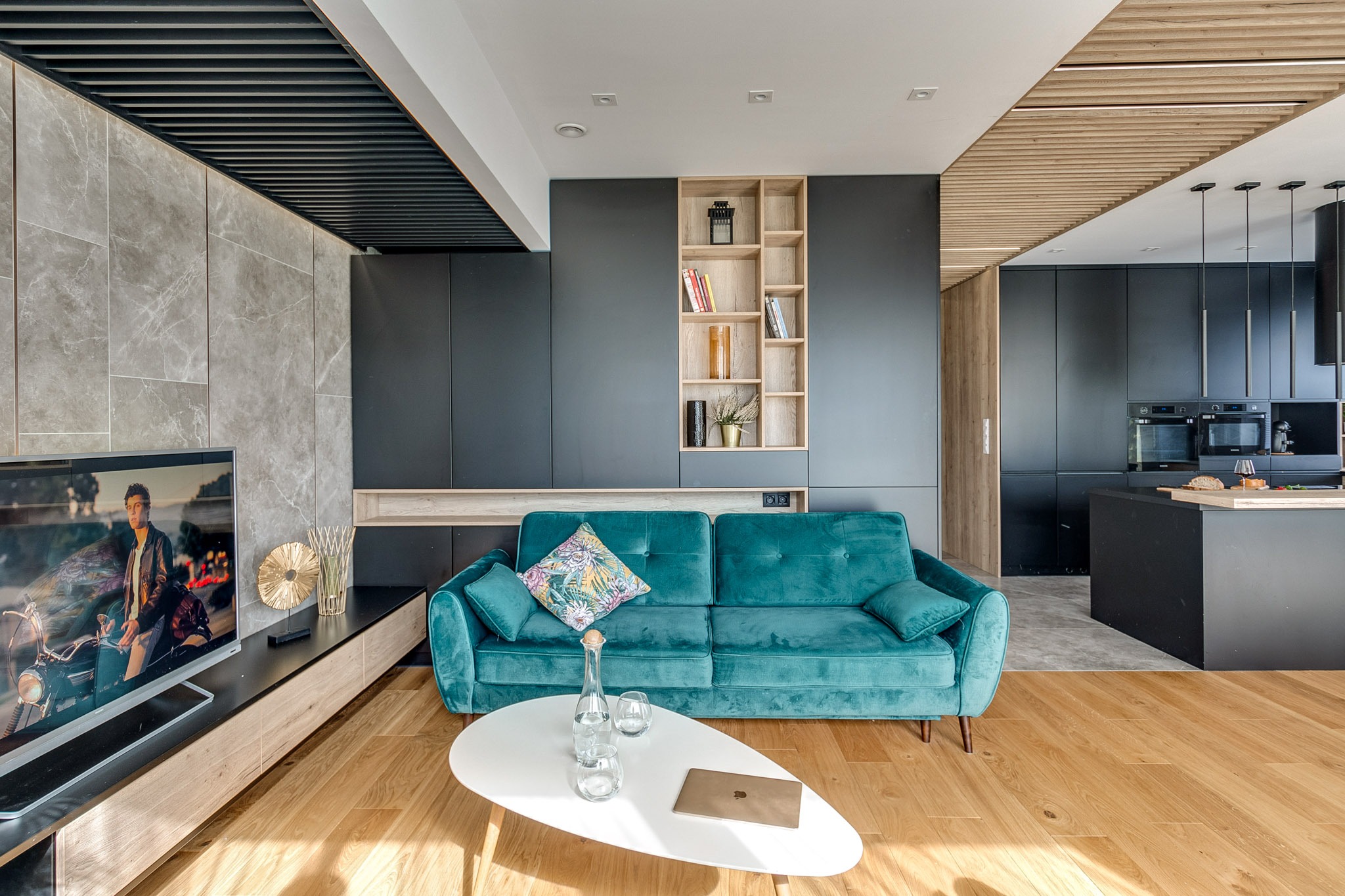 Nowoczesne wnętrze: turkusowa sofa, czarne szafki z półkami, drewniany sufit i podłoga. Na stole laptop i woda. Telewizor z filmem na ścianie z marmuru.