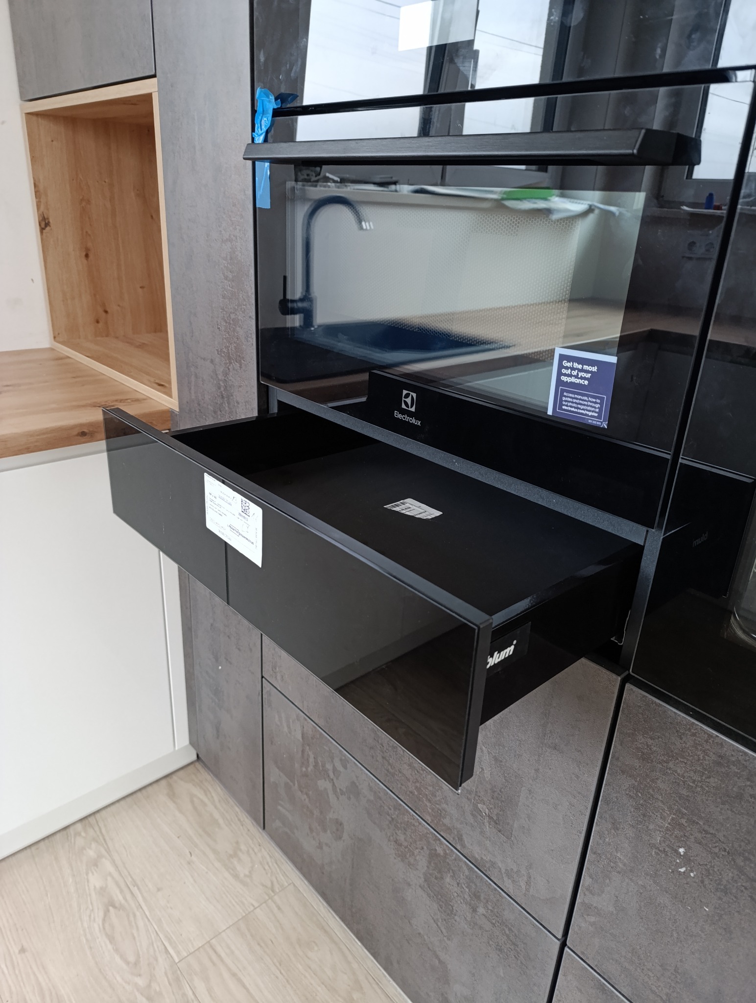 Częściowy widok nowoczesnej zabudowy kuchennej z otwartą, czarną szufladą z systemem Blum, fronty w odcieniach szarości, piekarnik Electrolux w tle.