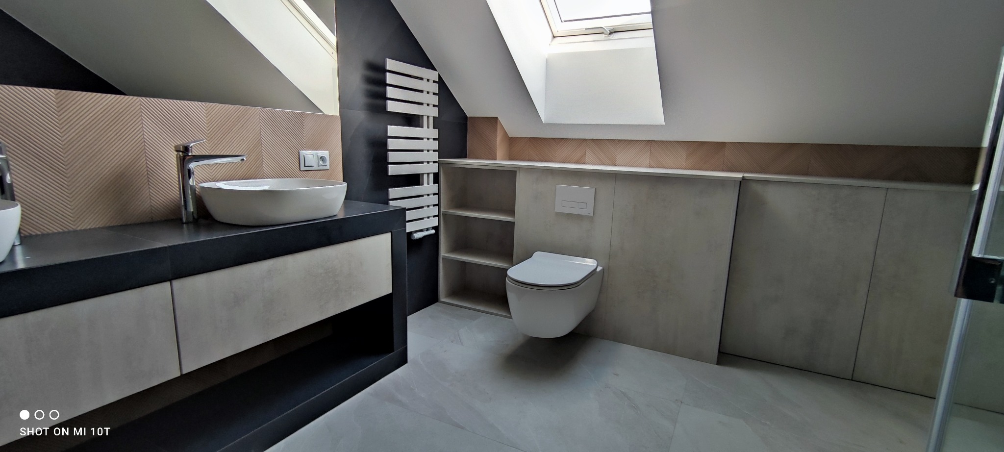 Nowoczesna łazienka na poddaszu z umywalką nablatową, grzejnikiem drabinkowym i zabudową WC w odcieniach szarości i czerni, oświetlona światłem z okna dachowego.