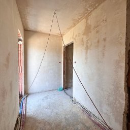 NTN CONSTRUCTION - Wnętrze w trakcie tynkowania: surowe ściany z widocznymi instalacjami elektrycznymi, fragment muru z cegły i linka pionująca zwisająca z sufitu.
