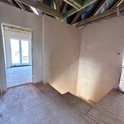 NTN CONSTRUCTION - Wnętrze domu w trakcie prac wykończeniowych: otynkowane ściany, surowy strop z drewnianych belek, widoczne schody i okno. Wnętrze w stanie deweloperskim.