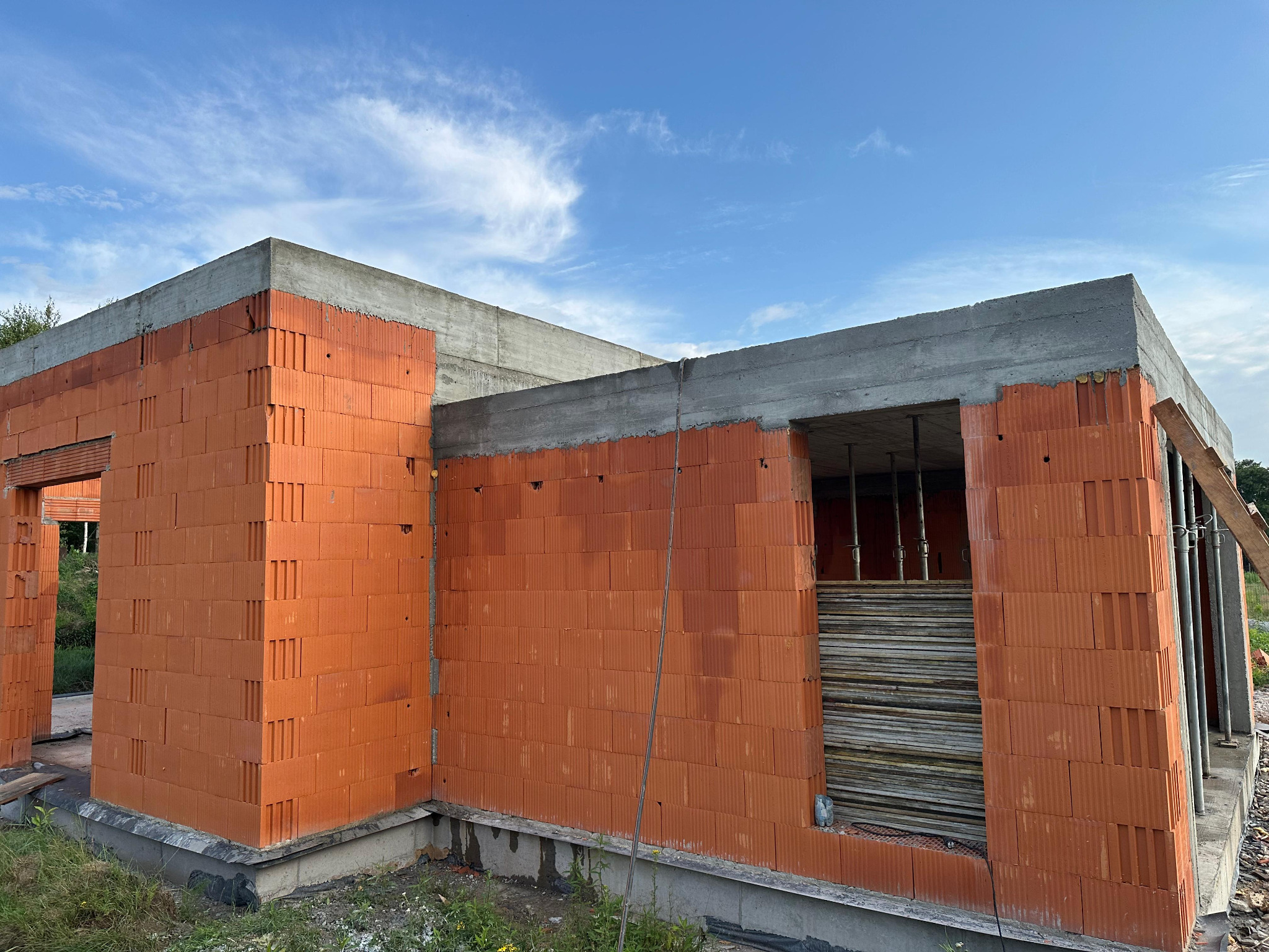 Surowy stan domu z czerwonej cegły ceramicznej, z widocznym betonowym wieńcem i podtrzymującymi stemplami w otworze okiennym, na tle błękitnego nieba.