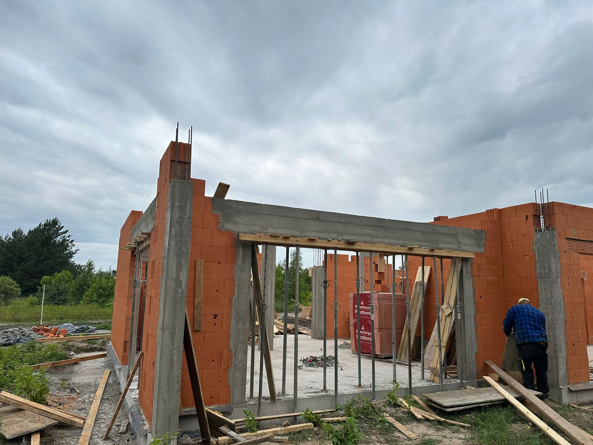 Budowa domu z czerwonej cegły: ściany wzniesione, betonowe nadproża, widoczne zbrojenia. W tle pracownik w niebieskiej koszuli. Pochmurne niebo.