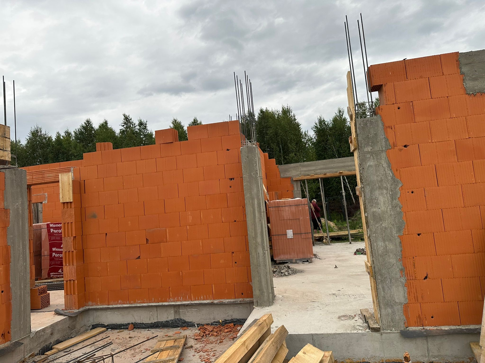 Wznoszenie ścian z czerwonej cegły Porotherm, widoczne zbrojenia i betonowe słupy. W tle zalesiony teren i pochmurne niebo.