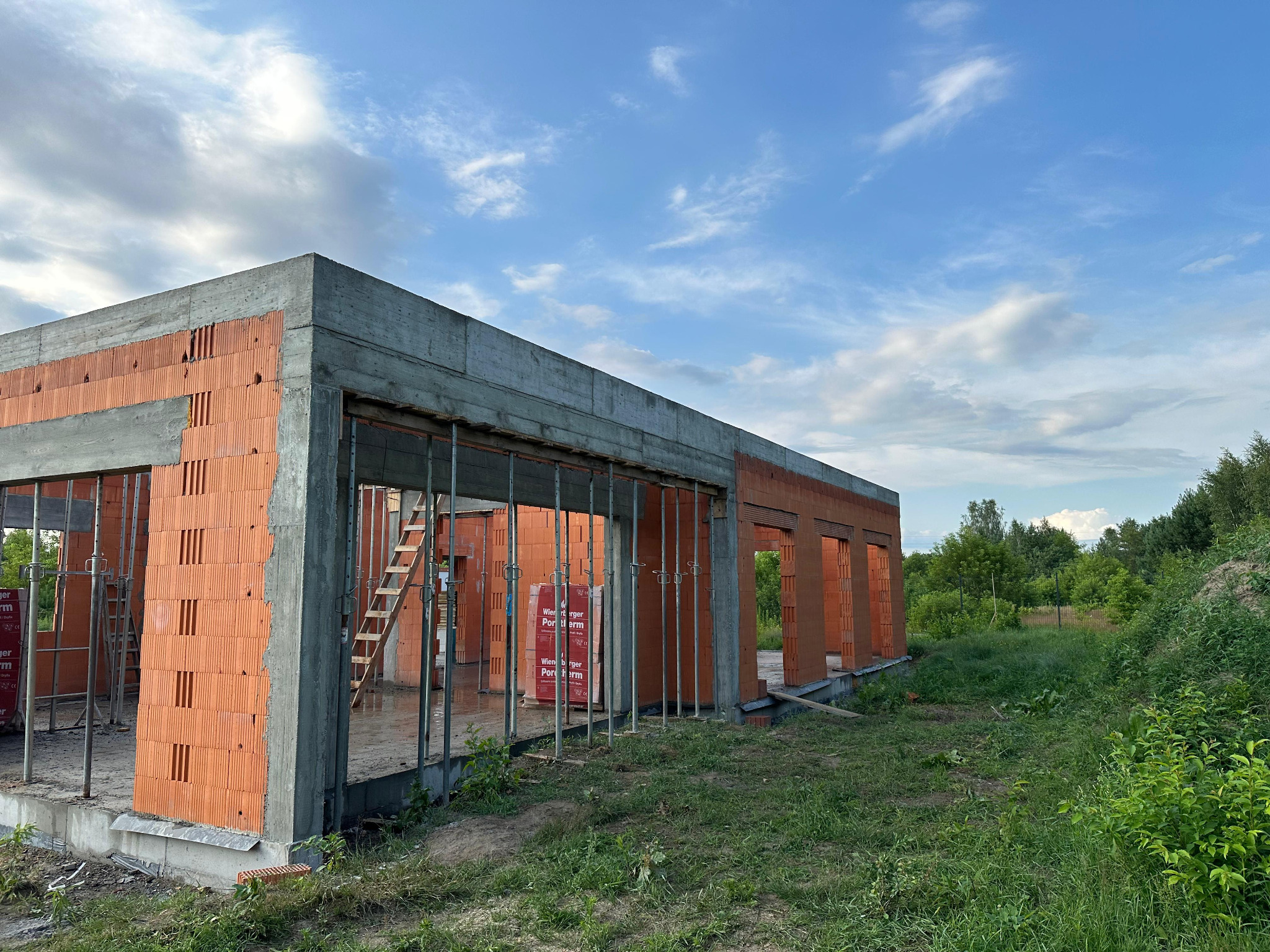 Surowy stan budynku z czerwonej cegły, z widocznymi betonowymi elementami konstrukcyjnymi i podporami, na tle błękitnego nieba z chmurami.