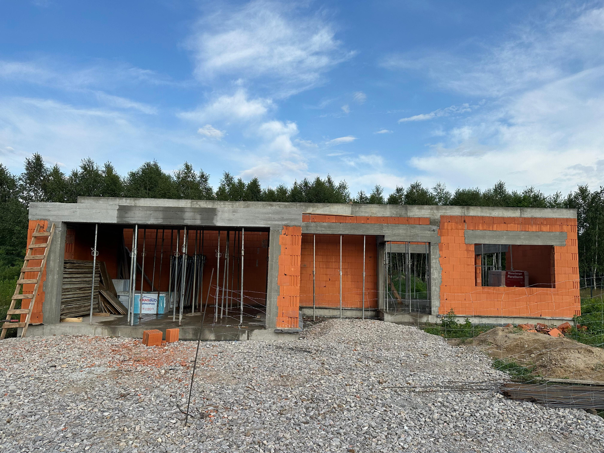 Surowy stan domu z czerwonej cegły, widoczne szalunki i podpory, na pierwszym planie gruz i zbrojenia, w tle zieleń drzew i błękitne niebo.