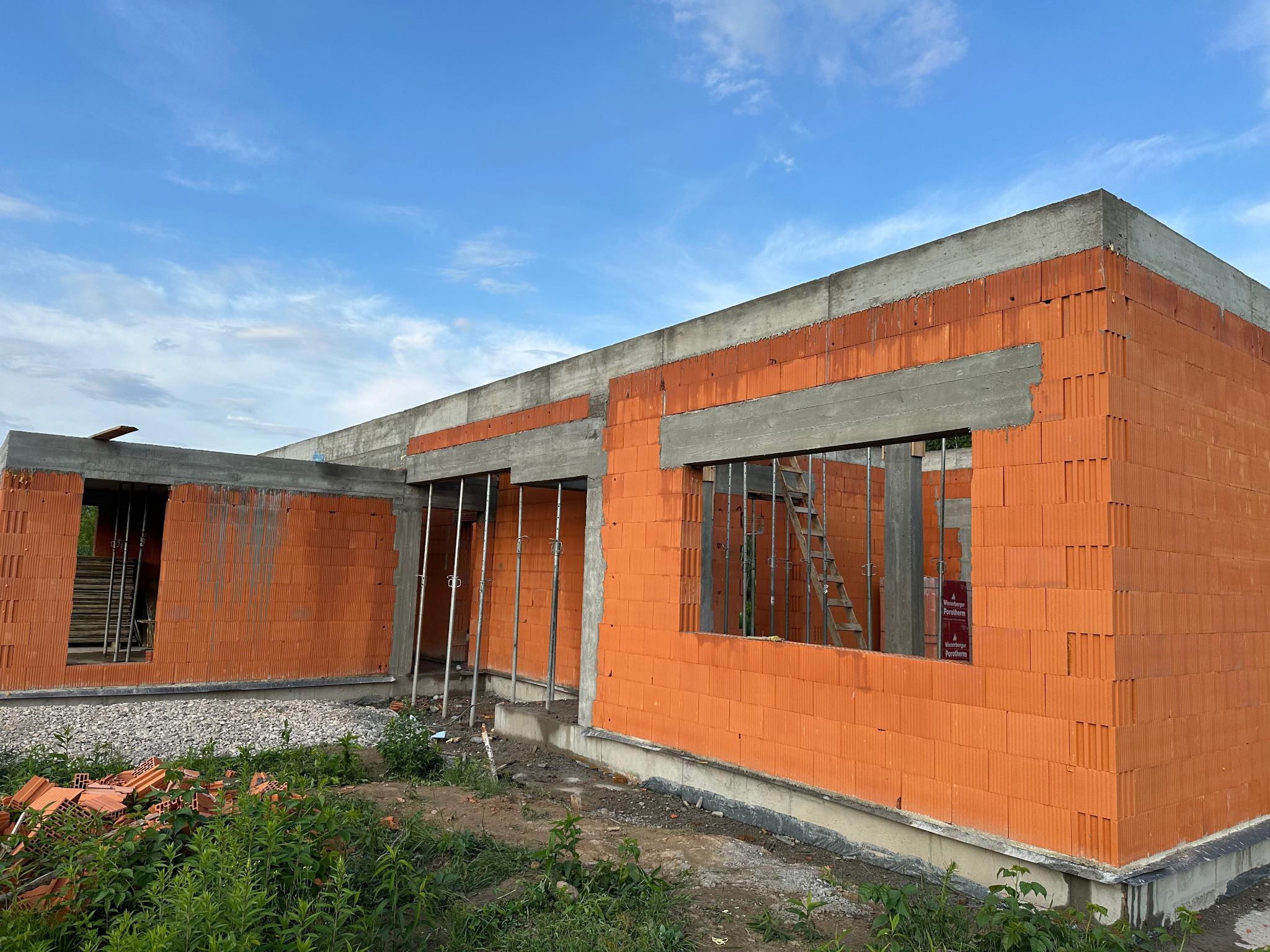 Niedokończony budynek z czerwonej cegły Porotherm z betonowymi nadprożami i słupami, widoczne podpory i drabina, na pierwszym planie gruz i roślinność.
