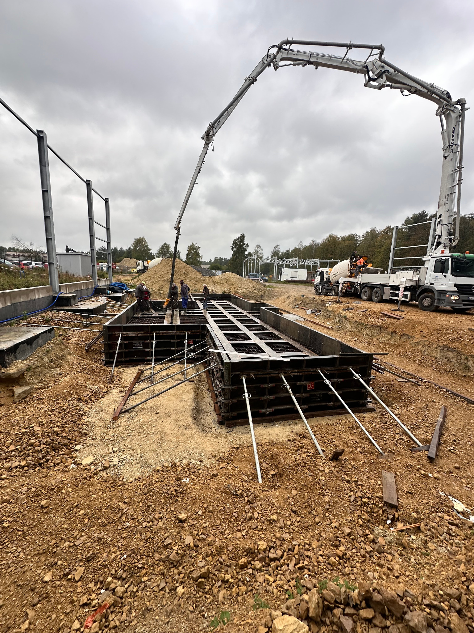 Wylewanie betonu do szalunków fundamentowych z użyciem pompy do betonu. Widoczni pracownicy w kaskach i kamizelkach odblaskowych. W tle ciężarówka z betonomieszarką.