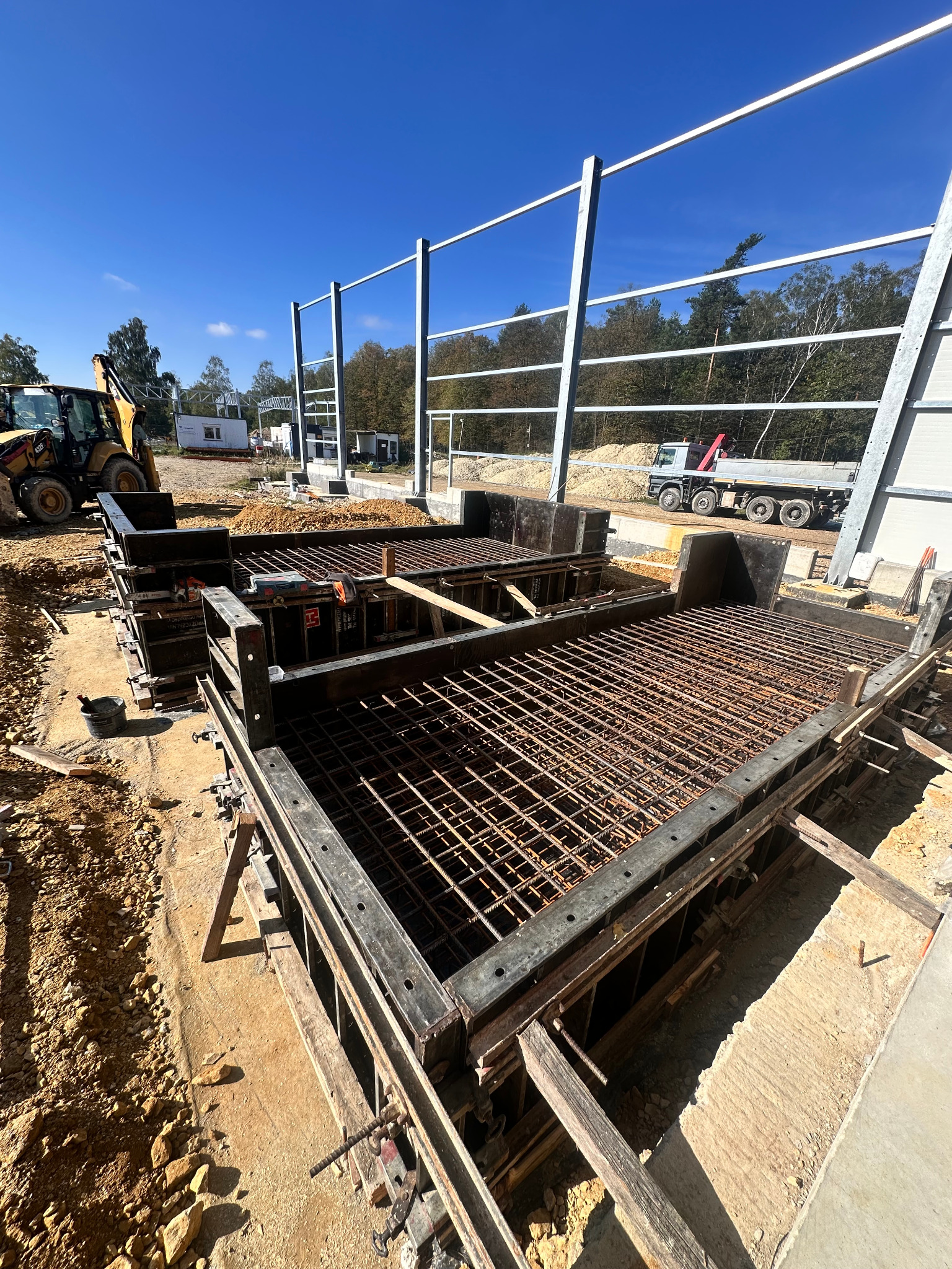Zbrojenie fundamentów pod budynek przemysłowy z użyciem szalunków systemowych. Na budowie widoczny sprzęt ciężki i konstrukcja stalowa budynku.