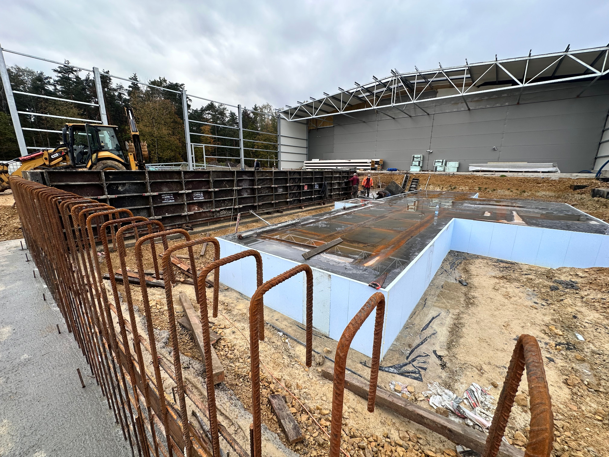 Fundamenty budynku przemysłowego w trakcie budowy: zbrojenie, szalunki i izolacja termiczna. W tle widoczny ciężki sprzęt budowlany i konstrukcja stalowa hali.