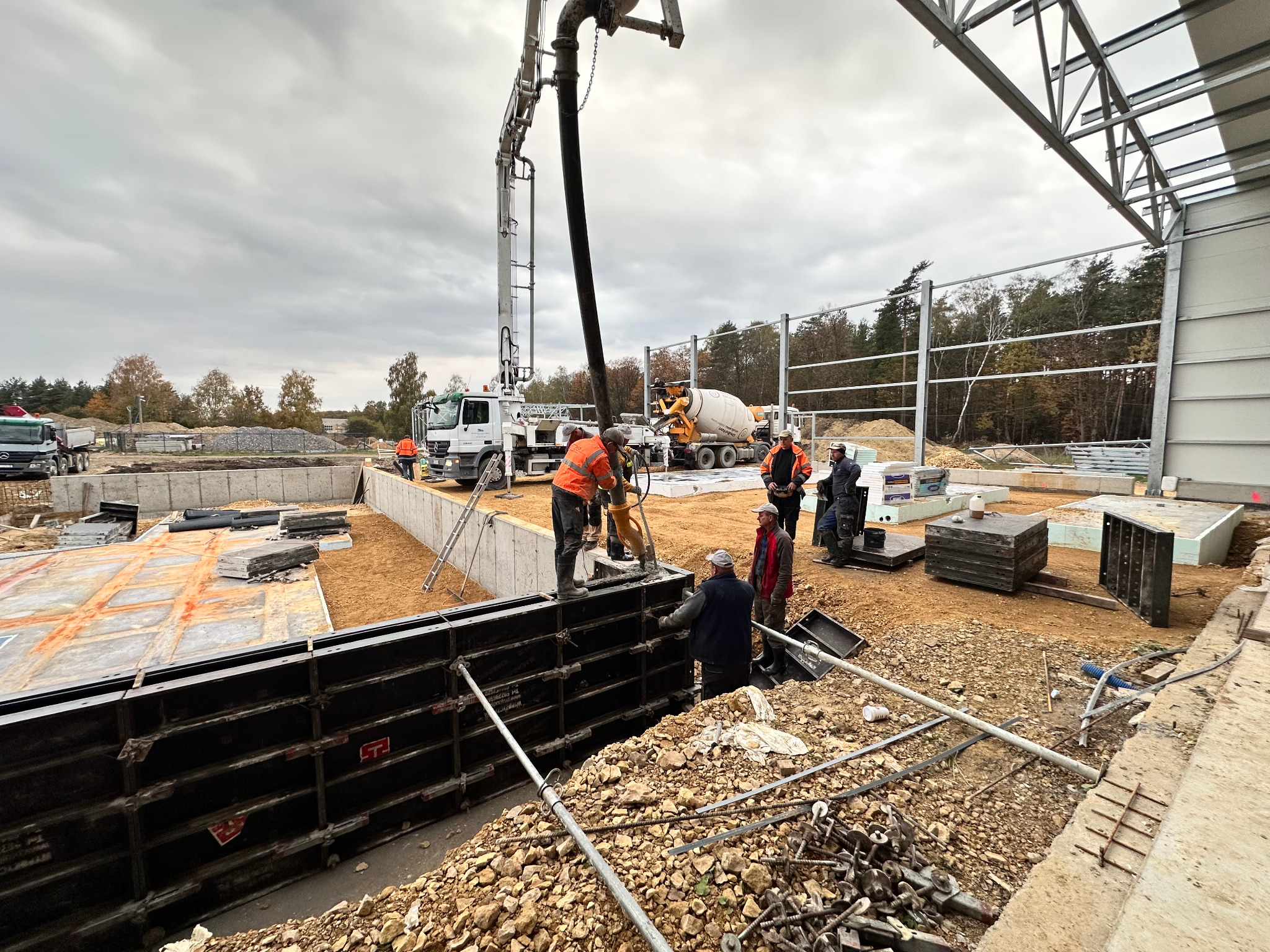 Wylewanie betonu do szalunków fundamentowych z użyciem pompy do betonu. Widoczni pracownicy w odblaskowych kamizelkach i ciężki sprzęt budowlany.