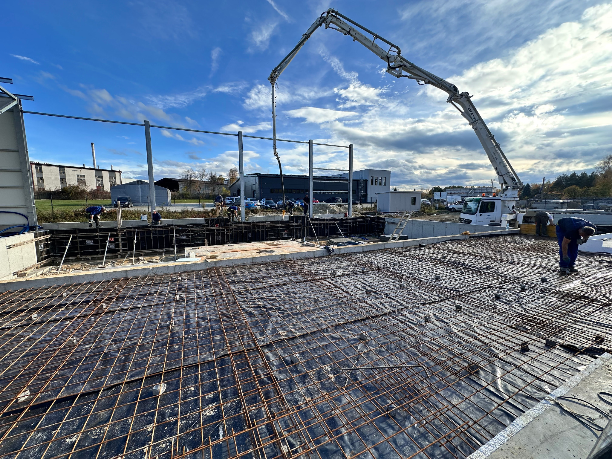 Zbrojenie fundamentów pod budynek z użyciem pompy do betonu. Widoczni pracownicy w trakcie wykonywania prac zbrojarskich. Błękitne niebo w tle.