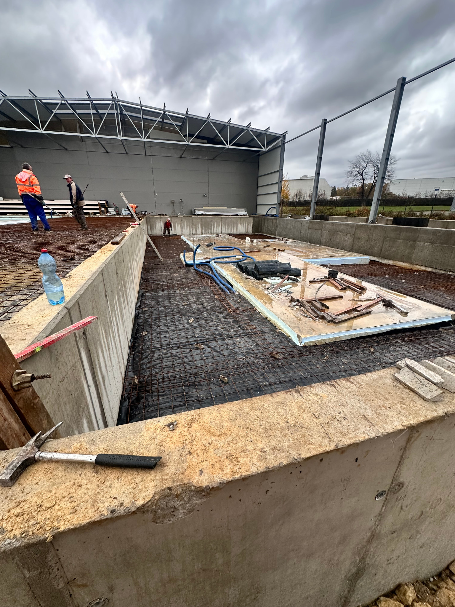Zbrojenie fundamentów pod budynek przemysłowy z widocznymi pracownikami w oddali i betonowymi ścianami szalunkowymi. Na pierwszym planie młotek.