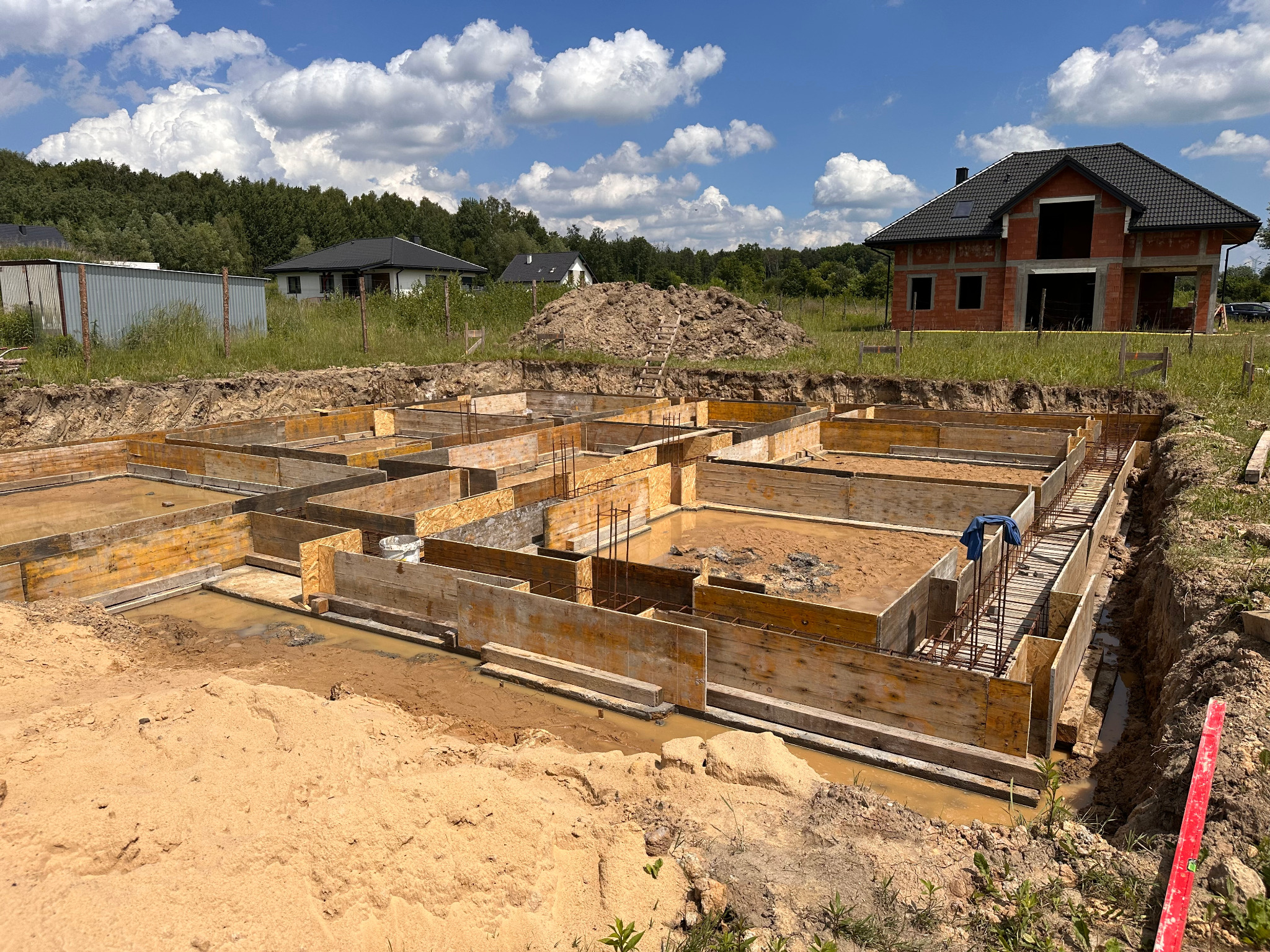 Szczegółowy widok wykopu z drewnianym szalunkiem i zbrojeniem pod fundamenty domu jednorodzinnego w trakcie budowy, z widocznym budynkiem w tle.