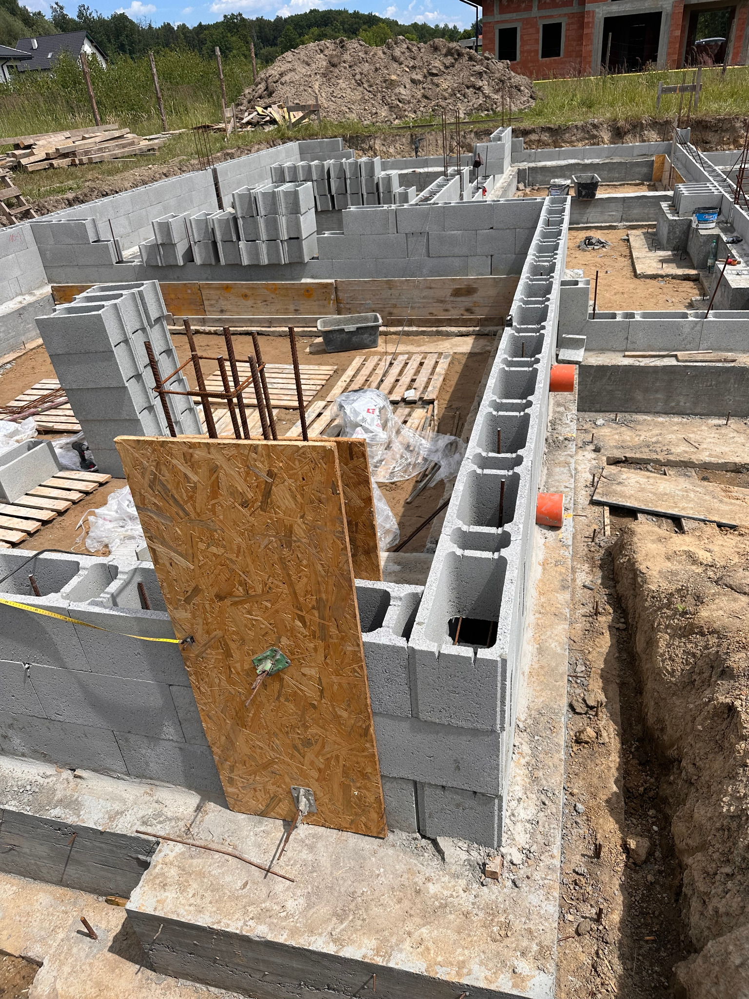 Wysoki kąt na budowę fundamentów z bloczków betonowych, z widocznymi deskami szalunkowymi i wystającymi prętami zbrojeniowymi. W tle hałda ziemi i fragment budynku w budowie.