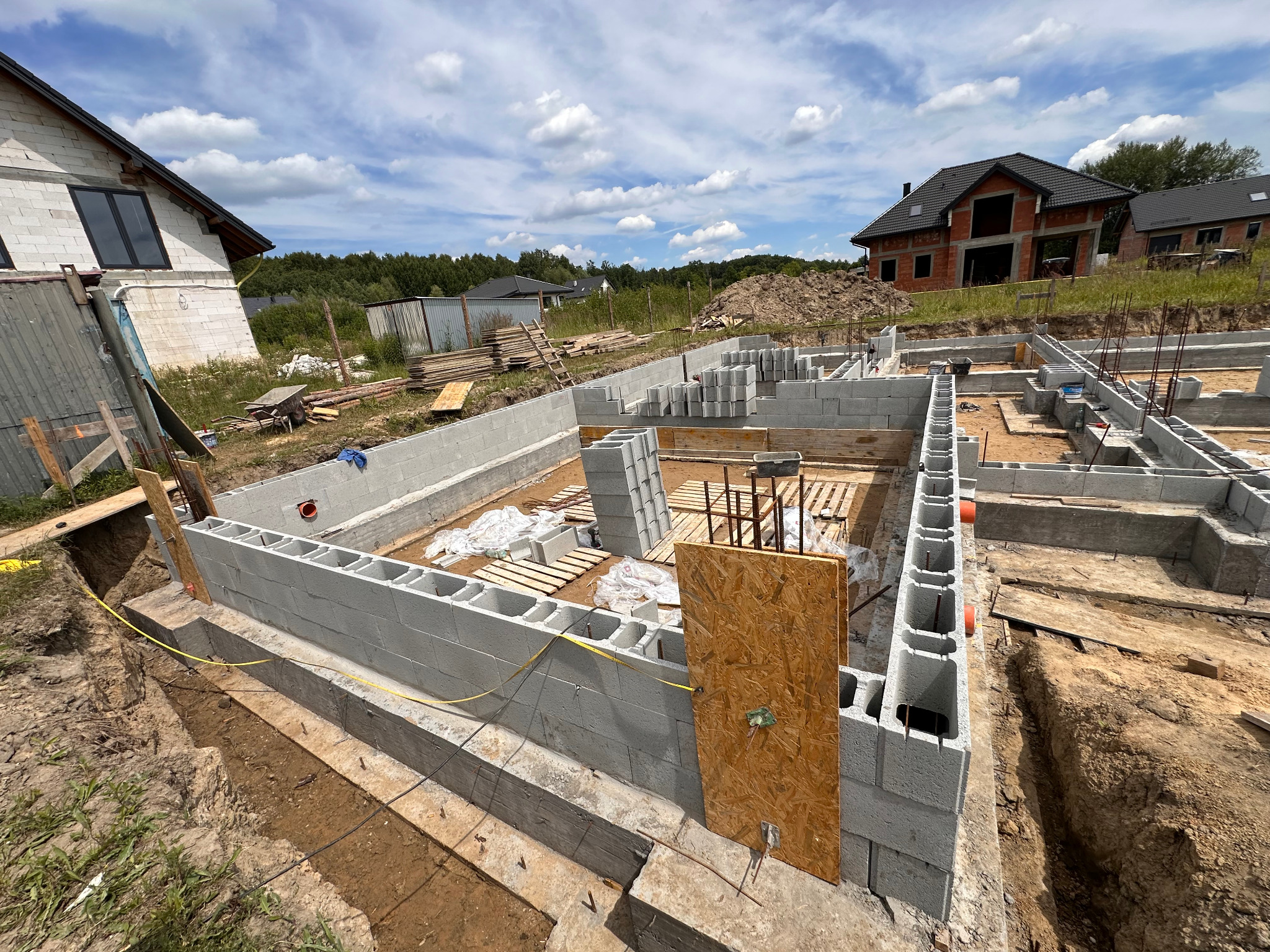 Fundamenty z bloczków betonowych na budowie domu jednorodzinnego, widoczne zbrojenia i wykop pod instalacje. W tle dom w stanie surowym otwartym.