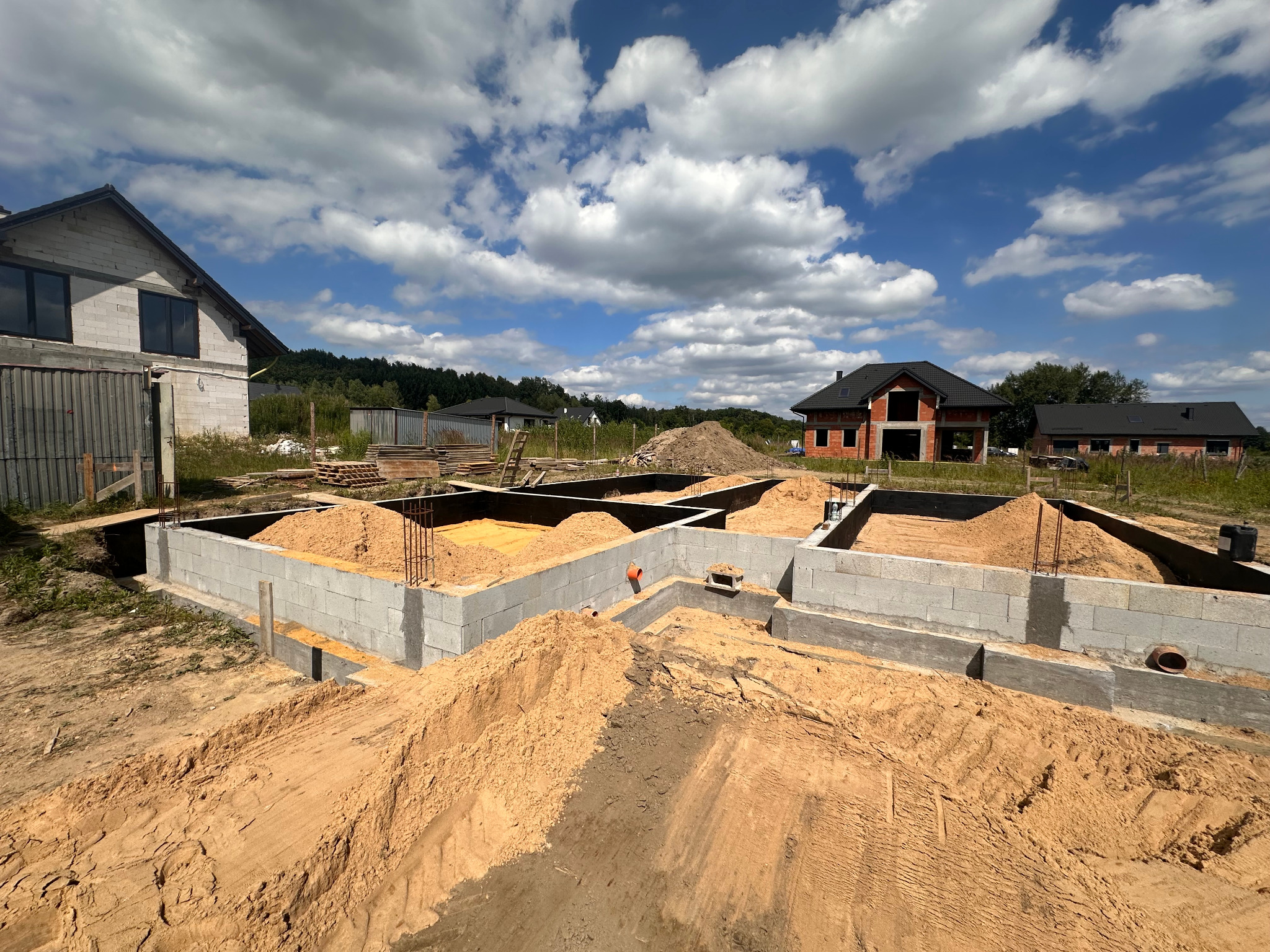 Wykopane fundamenty pod budowę domu, częściowo wypełnione piaskiem, z widocznymi zbrojeniami i elementami instalacji. W tle domy w różnym stanie budowy i błękitne niebo.