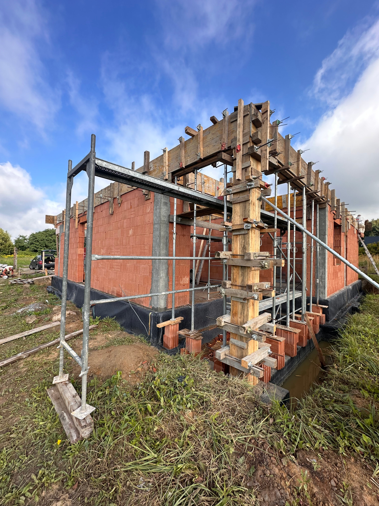 Budowa domu z czerwonej cegły, widoczne drewniane szalunki i rusztowania. Etap wznoszenia ścian i przygotowania do zalewania betonem. Błękitne niebo w tle.