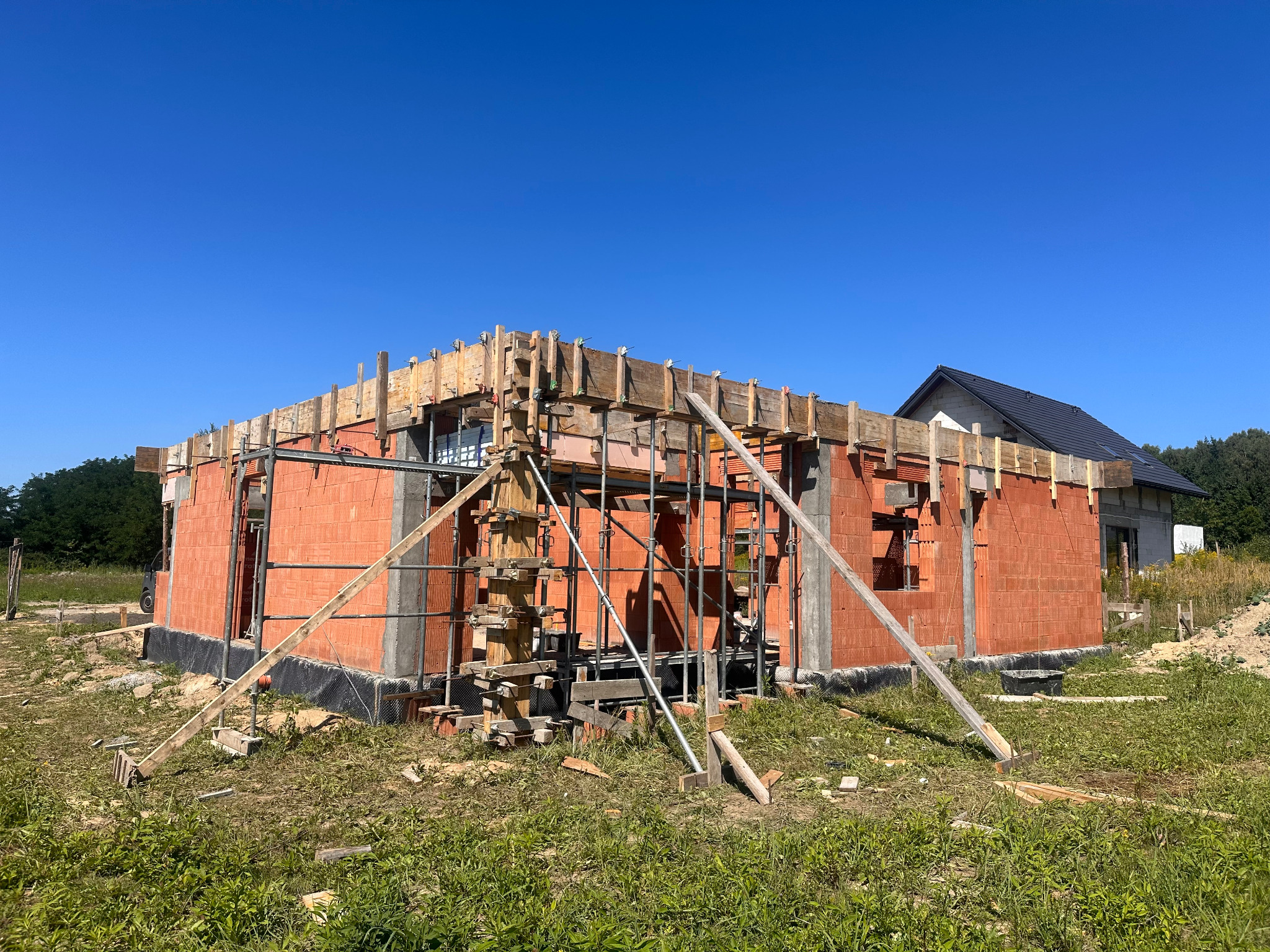 Ceglany dom w trakcie budowy, z drewnianym szalunkiem na wieńcu, na tle błękitnego nieba i zieleni. Widoczny fragment dachu sąsiedniego budynku.