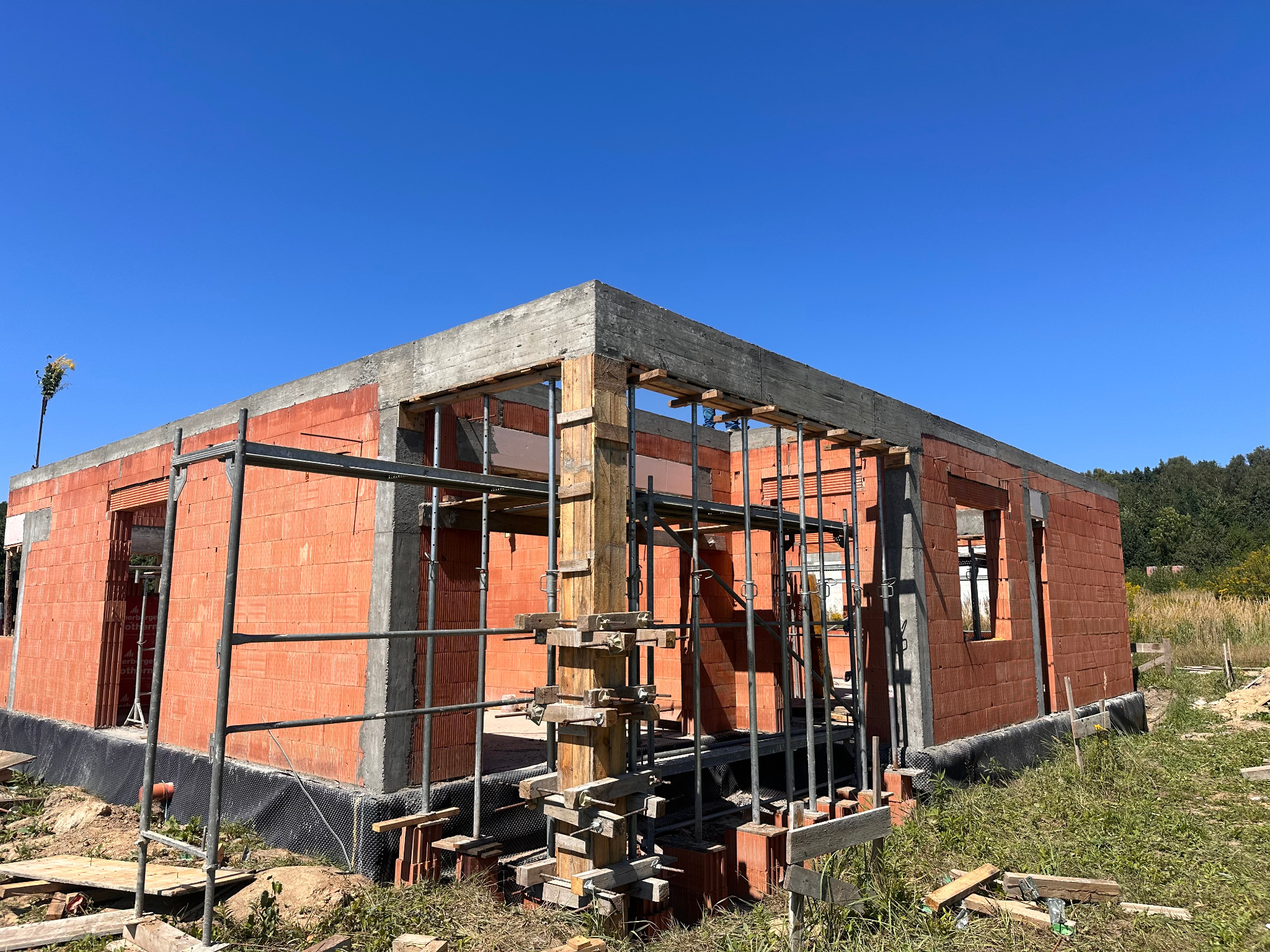 Budowa domu jednorodzinnego z czerwonej cegły, widoczne szalunki i zbrojenia, w tle błękitne niebo i zieleń. Etap wznoszenia ścian i stropu.