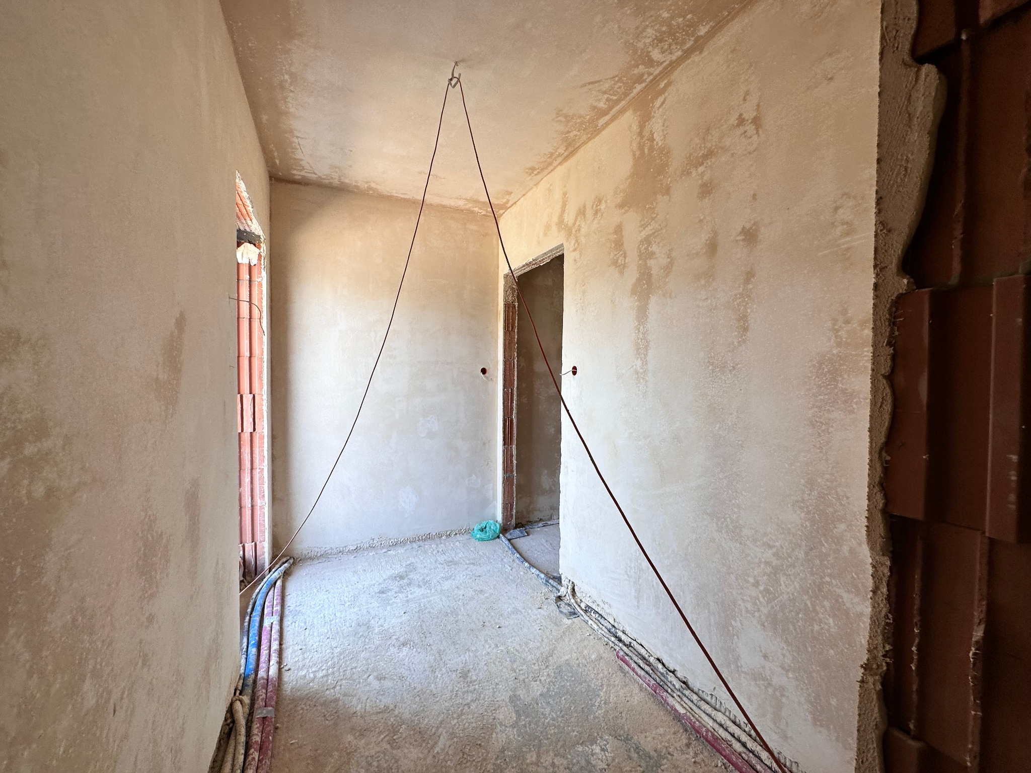 Wnętrze w trakcie tynkowania: surowe ściany z widocznymi instalacjami elektrycznymi, fragment muru z cegły i linka pionująca zwisająca z sufitu.