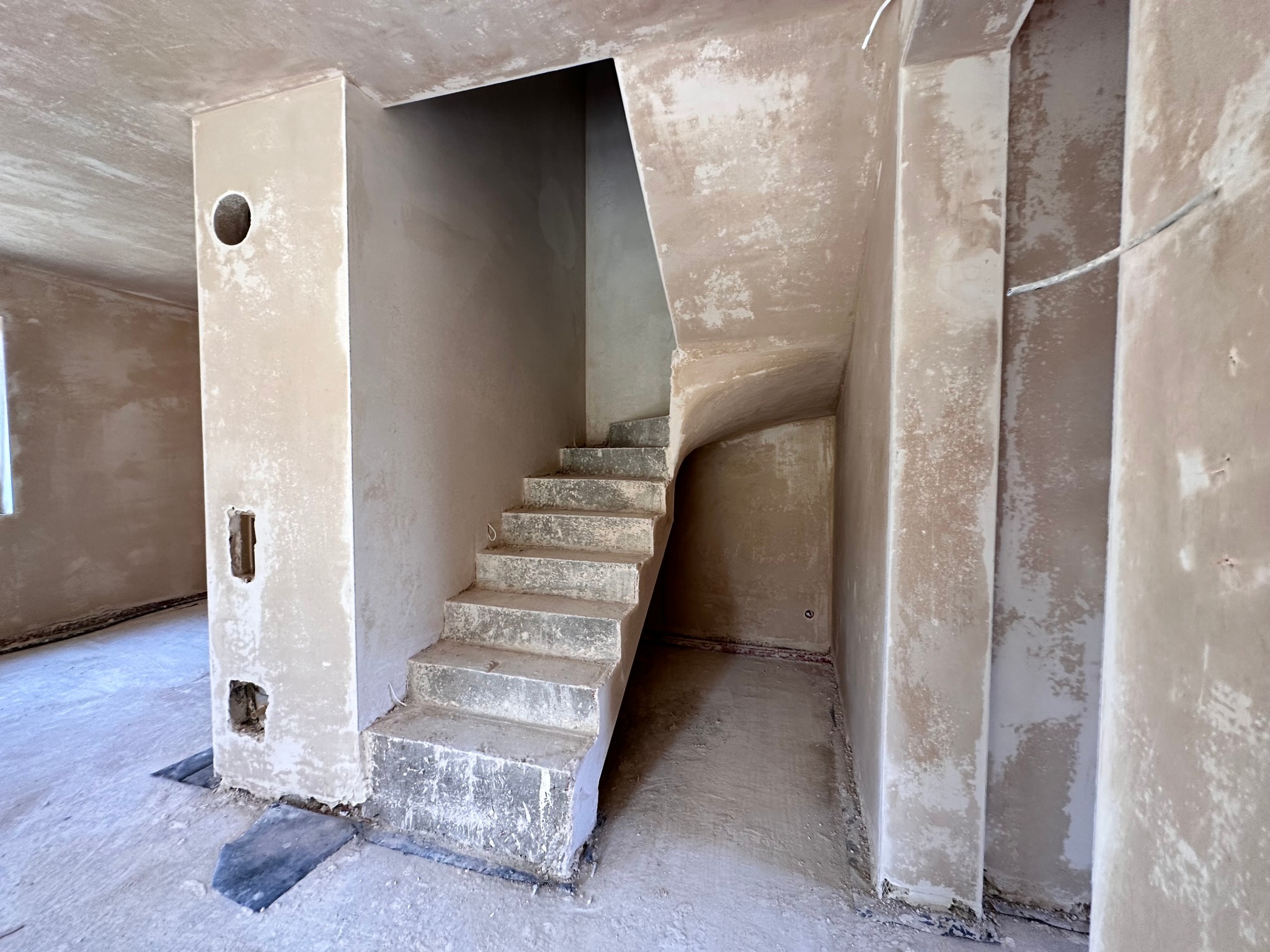 Surowe schody betonowe wewnątrz budynku w trakcie prac wykończeniowych, ściany pokryte tynkiem, widoczne otwory instalacyjne w filarze. Minimalistyczne ujęcie.