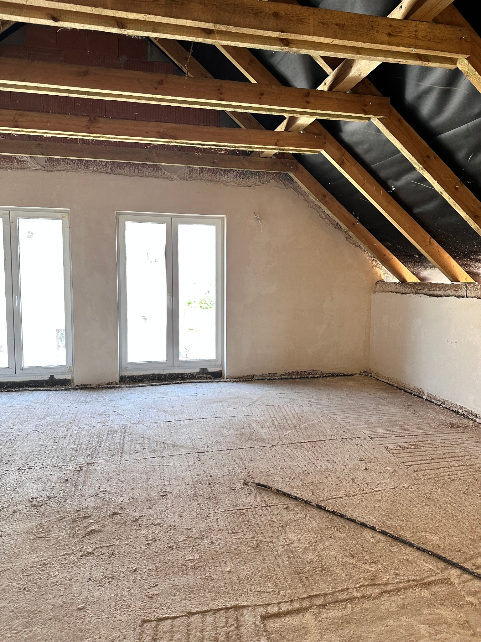 Poddasze z drewnianą konstrukcją dachu i oknami, ściany przygotowane do tynkowania, podłoga z wylewką. Widoczne ślady po instalacjach.