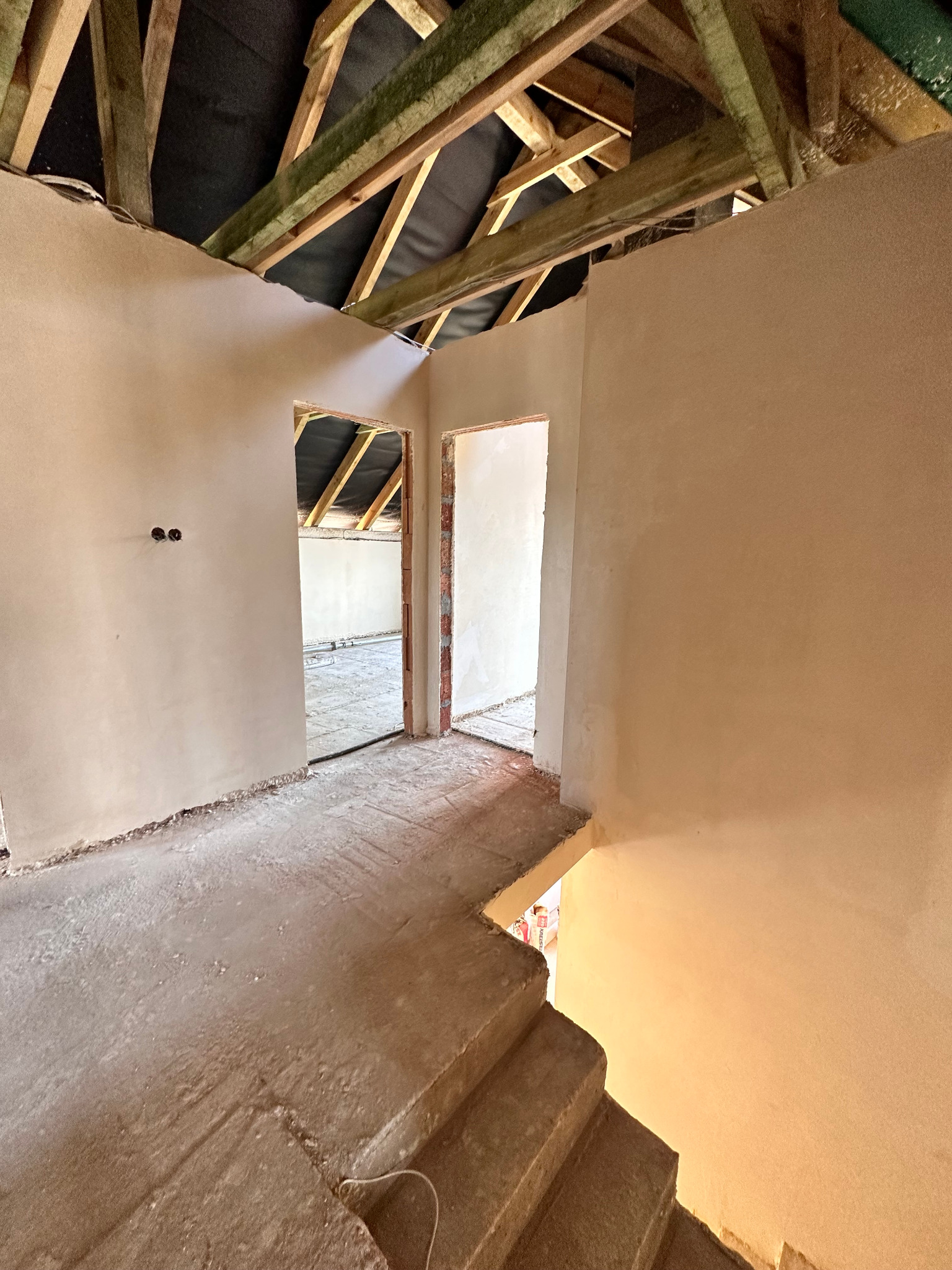 Wnętrze w stanie surowym: otynkowane ściany, drewniana konstrukcja dachu widoczna przez otwory drzwiowe. Schody prowadzą w dół, widoczne gniazdka elektryczne.