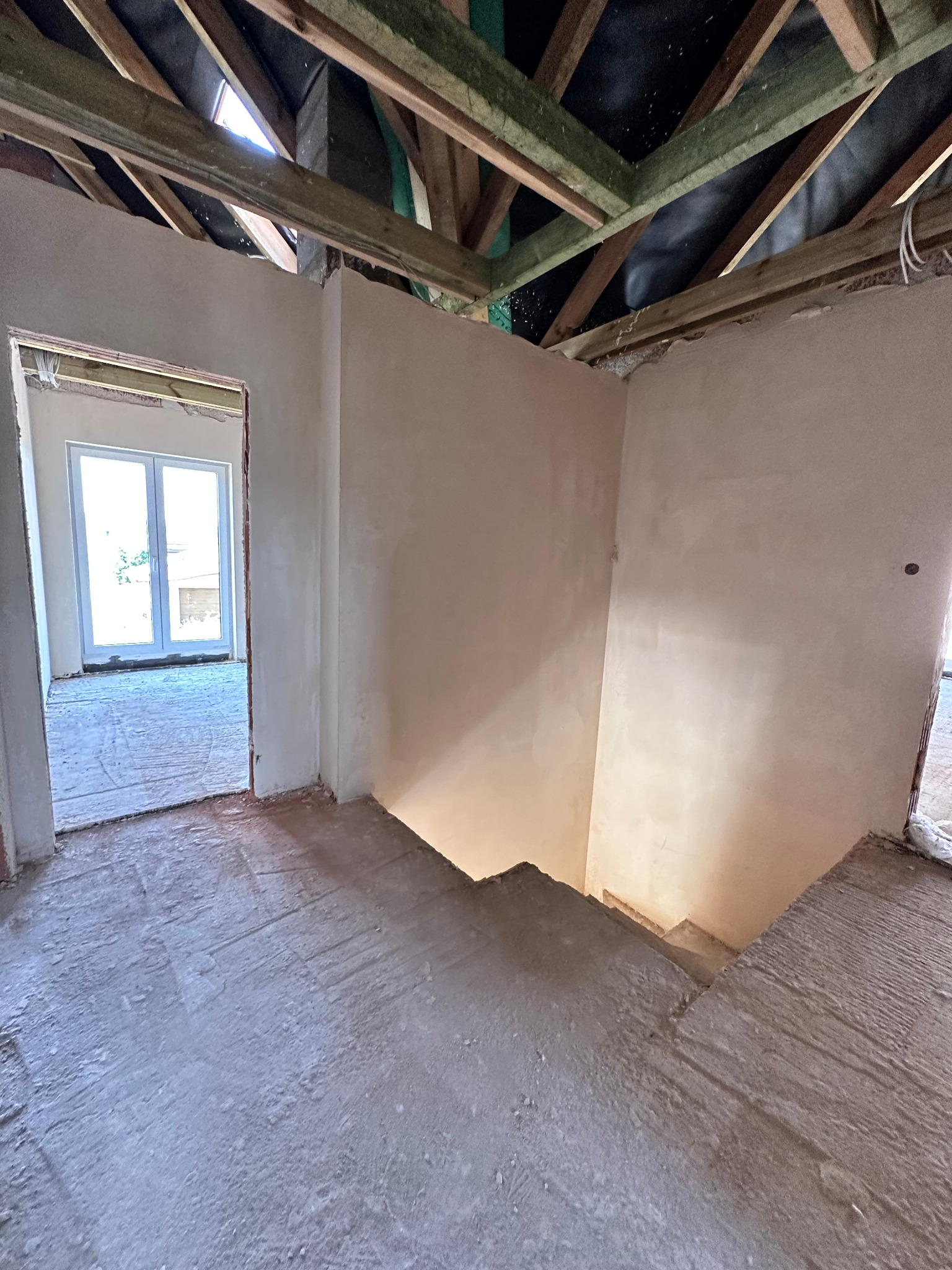 Wnętrze domu w trakcie prac wykończeniowych: otynkowane ściany, surowy strop z drewnianych belek, widoczne schody i okno. Wnętrze w stanie deweloperskim.