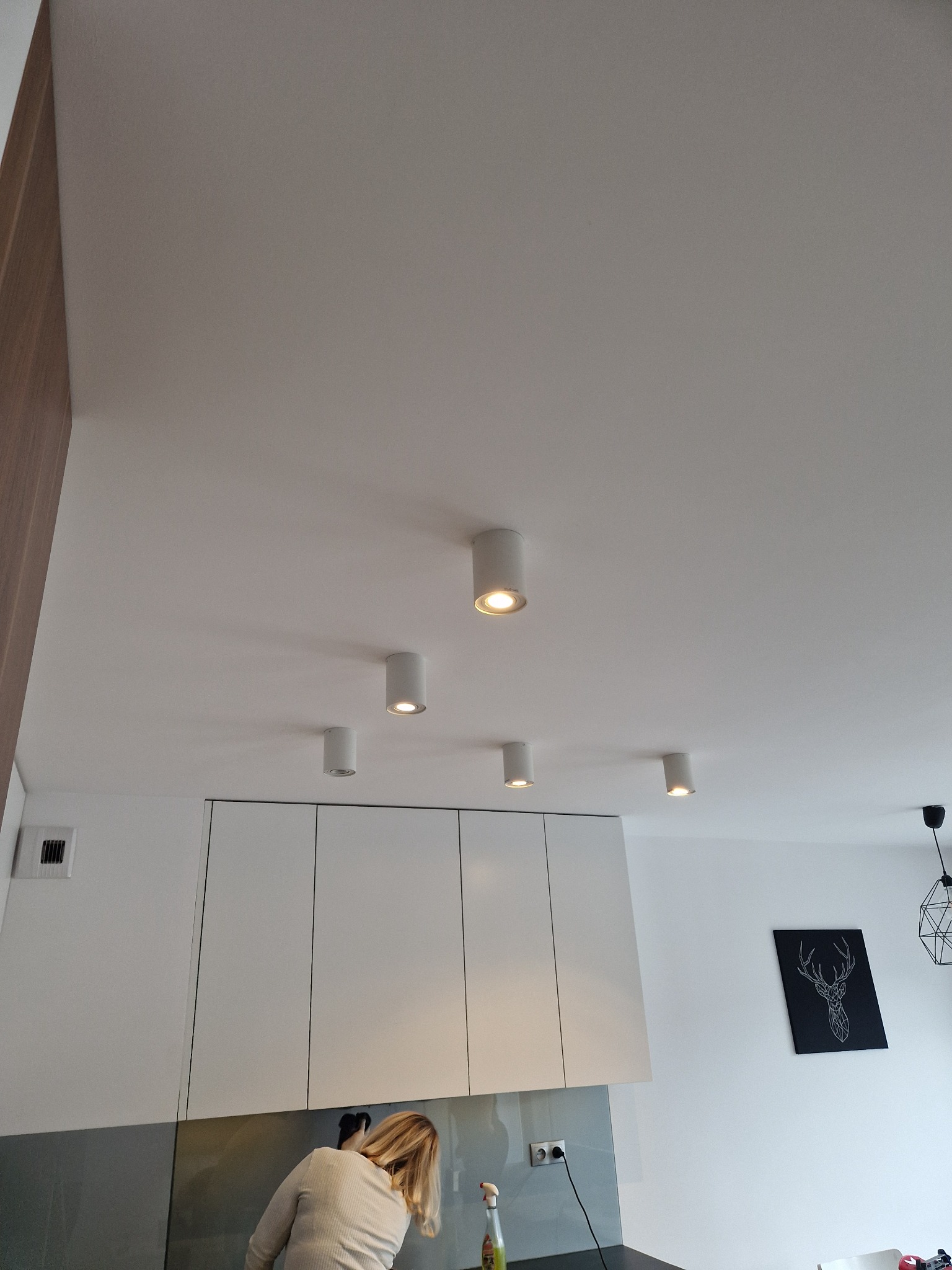 Białe, cylindryczne oprawy oświetleniowe LED zamontowane w suficie podwieszanym w nowoczesnej kuchni z białymi szafkami i szarą ścianą, widoczna osoba w tle oraz gniazdko elektryczne na ścianie.