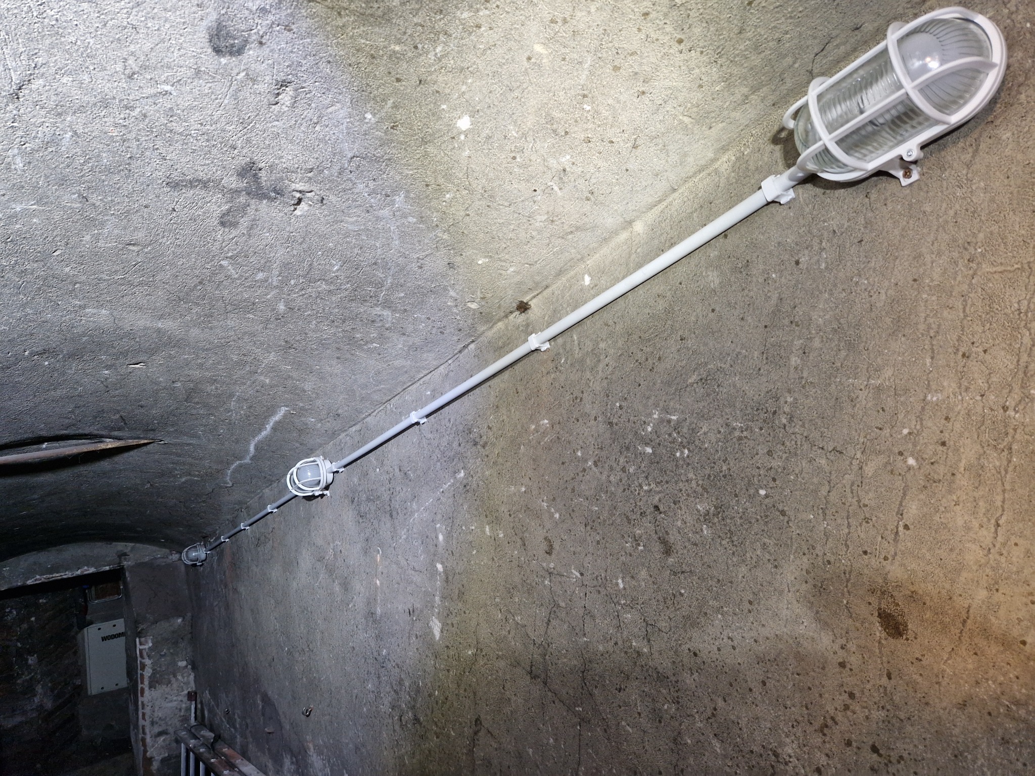 Oświetlenie korytarza piwnicznego z lampą w metalowej obudowie ochronnej, rurą instalacyjną i puszkami połączeniowymi, zamontowane na szorstkiej, betonowej ścianie i suficie.