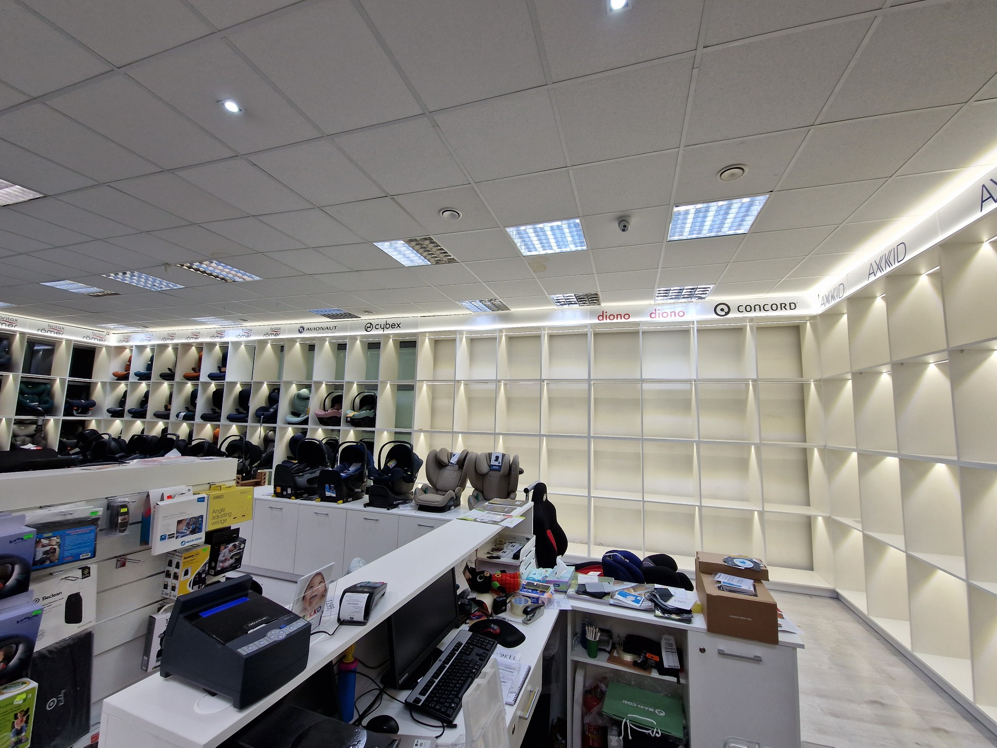 Wnętrze sklepu z fotelikami samochodowymi różnych marek, widok na regały z ekspozycją produktów i stanowisko obsługi klienta z komputerem i drukarką fiskalną.