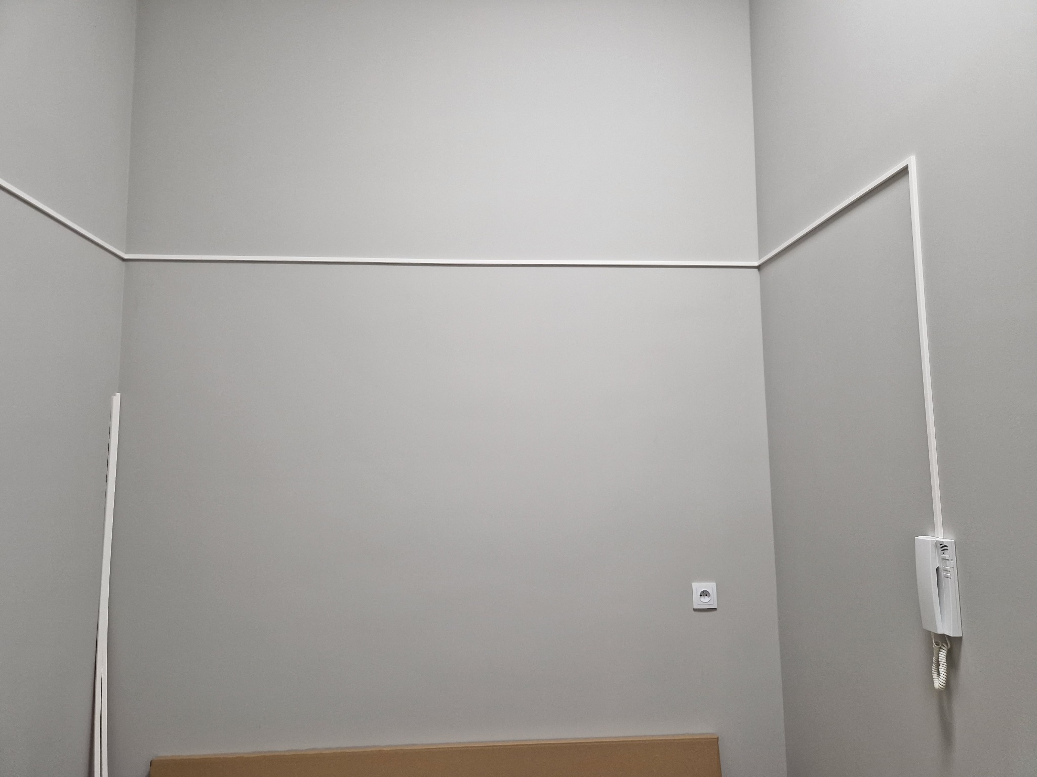 Szary pokój z zamontowanym domofonem na ścianie, białą listwą przypodłogową i gniazdkiem elektrycznym.