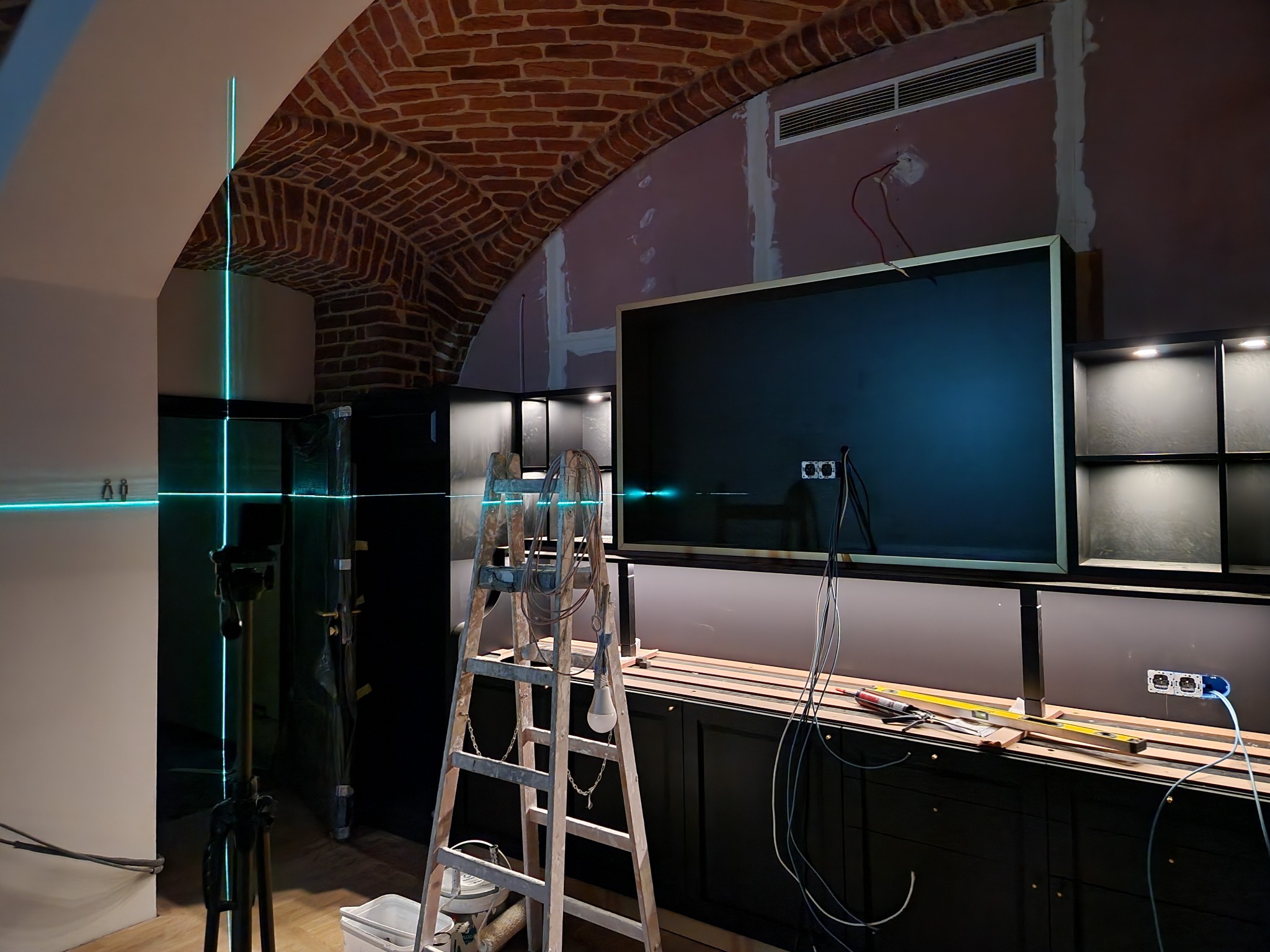 Wnętrze w trakcie prac wykończeniowych z widocznym laserowym niwelatorem ustawionym na statywie, wyznaczającym pion i poziom na tle surowego muru z cegły i przygotowanej podłoża pod instalację...