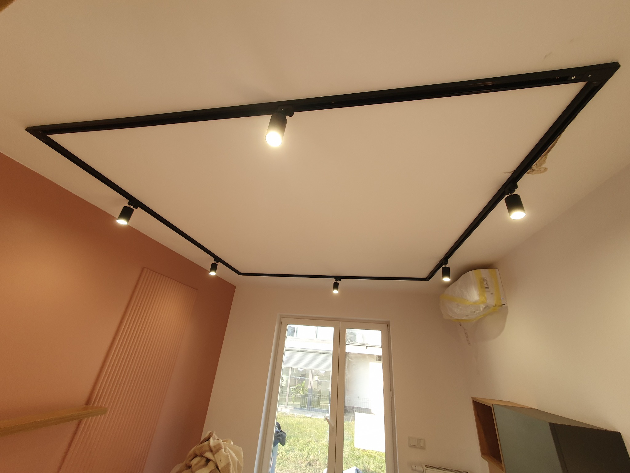 Czarna, kwadratowa szyna oświetleniowa sufitowa z reflektorami punktowymi, zamontowana w nowoczesnym wnętrzu z pomarańczową ścianą i oknem.