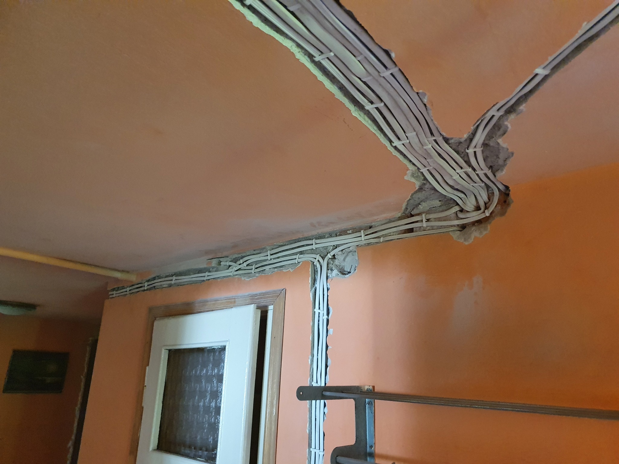 Rozprowadzenie przewodów elektrycznych w bruzdach na ścianie i suficie w pomarańczowym pomieszczeniu podczas remontu, widoczne białe kable zabezpieczone plastikowymi uchwytami.