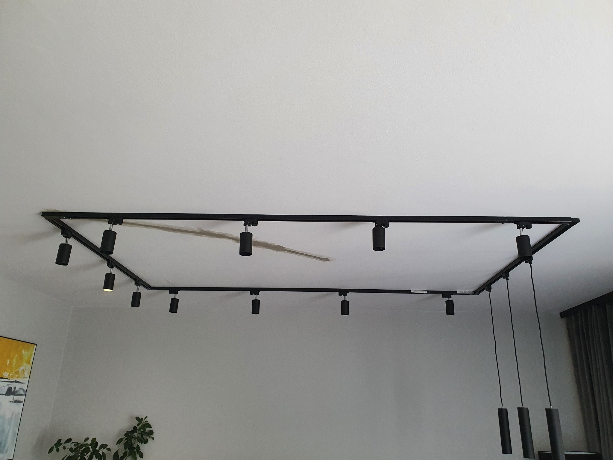 Czarna, kwadratowa szyna oświetleniowa z reflektorami punktowymi i trzema zwisającymi lampami cylindrycznymi, zamontowana na białym suficie w nowoczesnym wnętrzu z rośliną i obrazem w tle.