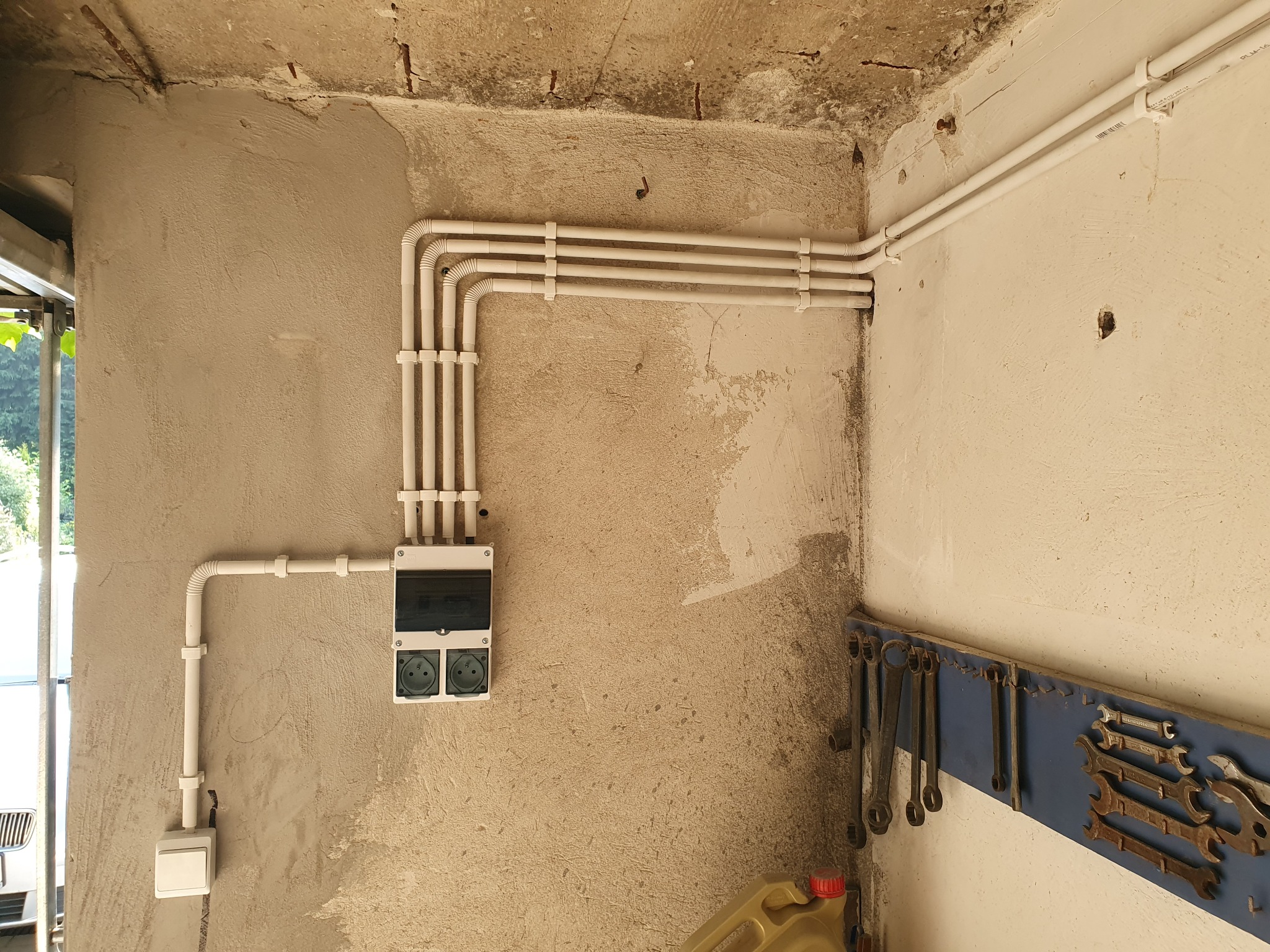 Białe kable elektryczne ułożone w równoległe rzędy na surowej, szarej ścianie garażu, z widoczną skrzynką z gniazdkami i włącznikiem, oraz wiszące klucze na niebieskiej tablicy w tle.