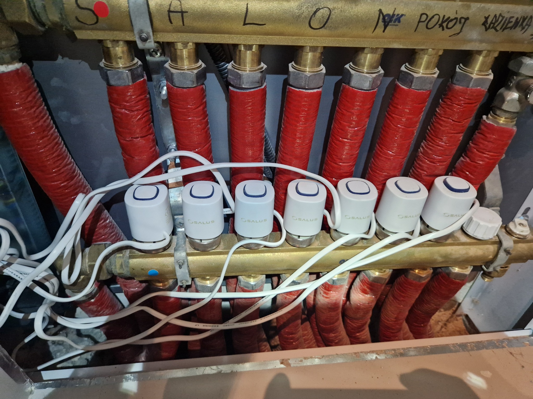 Rozdzielacz ogrzewania podłogowego z zamontowanymi, białymi głowicami termostatycznymi Salus, rury w izolacji termicznej, widoczne przewody elektryczne, napisy na rozdzielaczu.