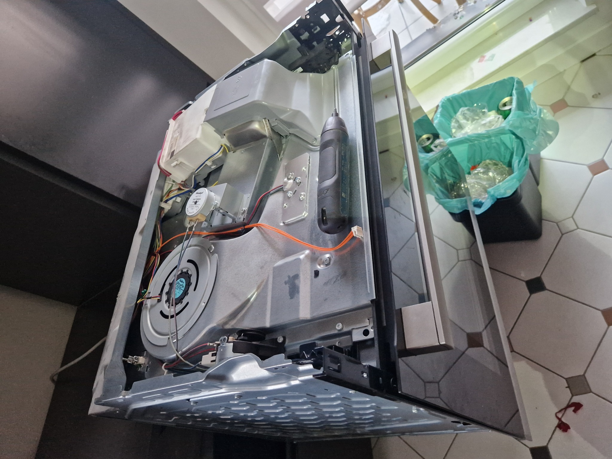 Wnętrze piekarnika elektrycznego z widocznymi elementami grzejnymi, wentylatorem i okablowaniem, obok śmietnik z puszkami.