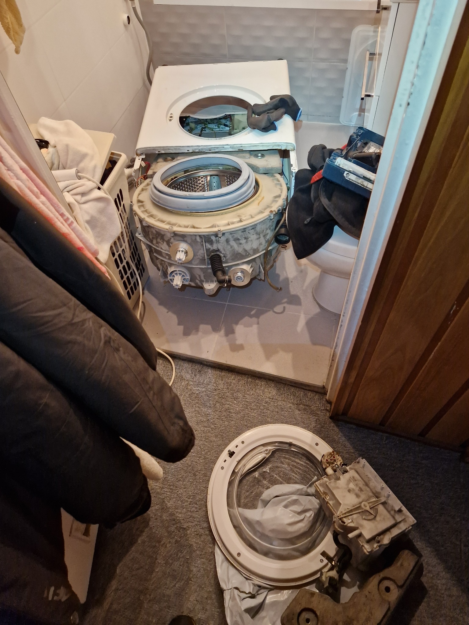 Rozłożona pralka w łazience podczas naprawy, widoczne elementy wewnętrzne i obudowa, narzędzia obok.