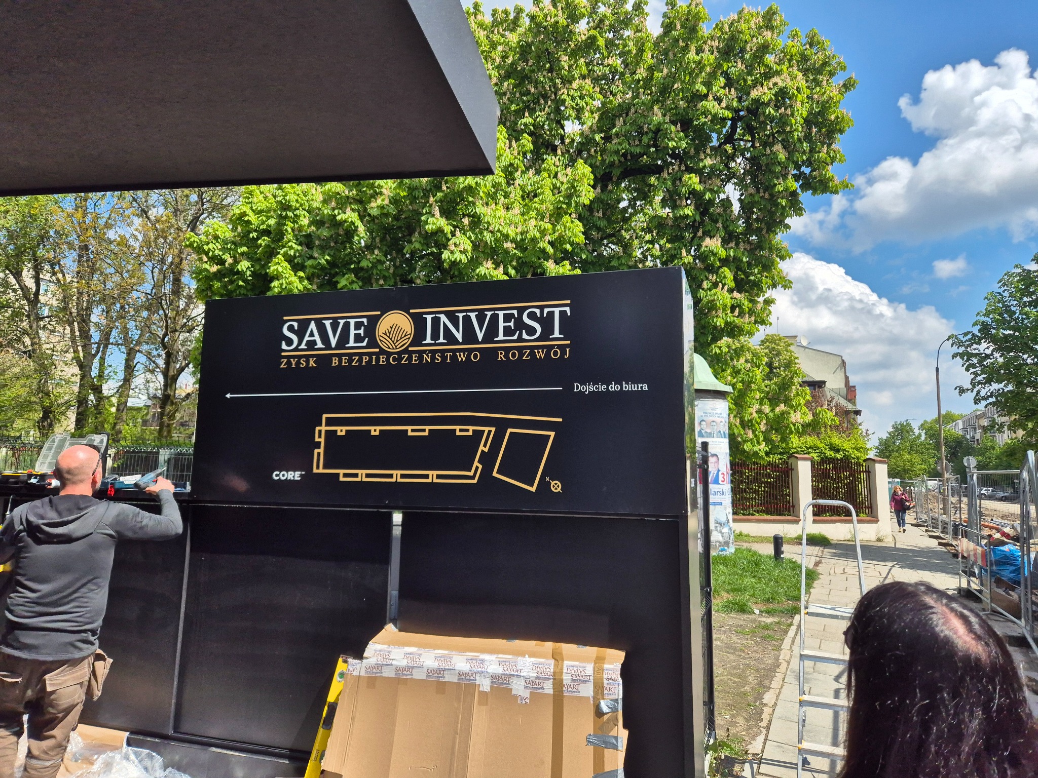 Pracownik montuje czarny, podświetlany szyld reklamowy z napisem 'SAVE INVEST' i planem biura, widoczne narzędzia i kartony zabezpieczające, w tle zieleń miejska i fragment chodnika.