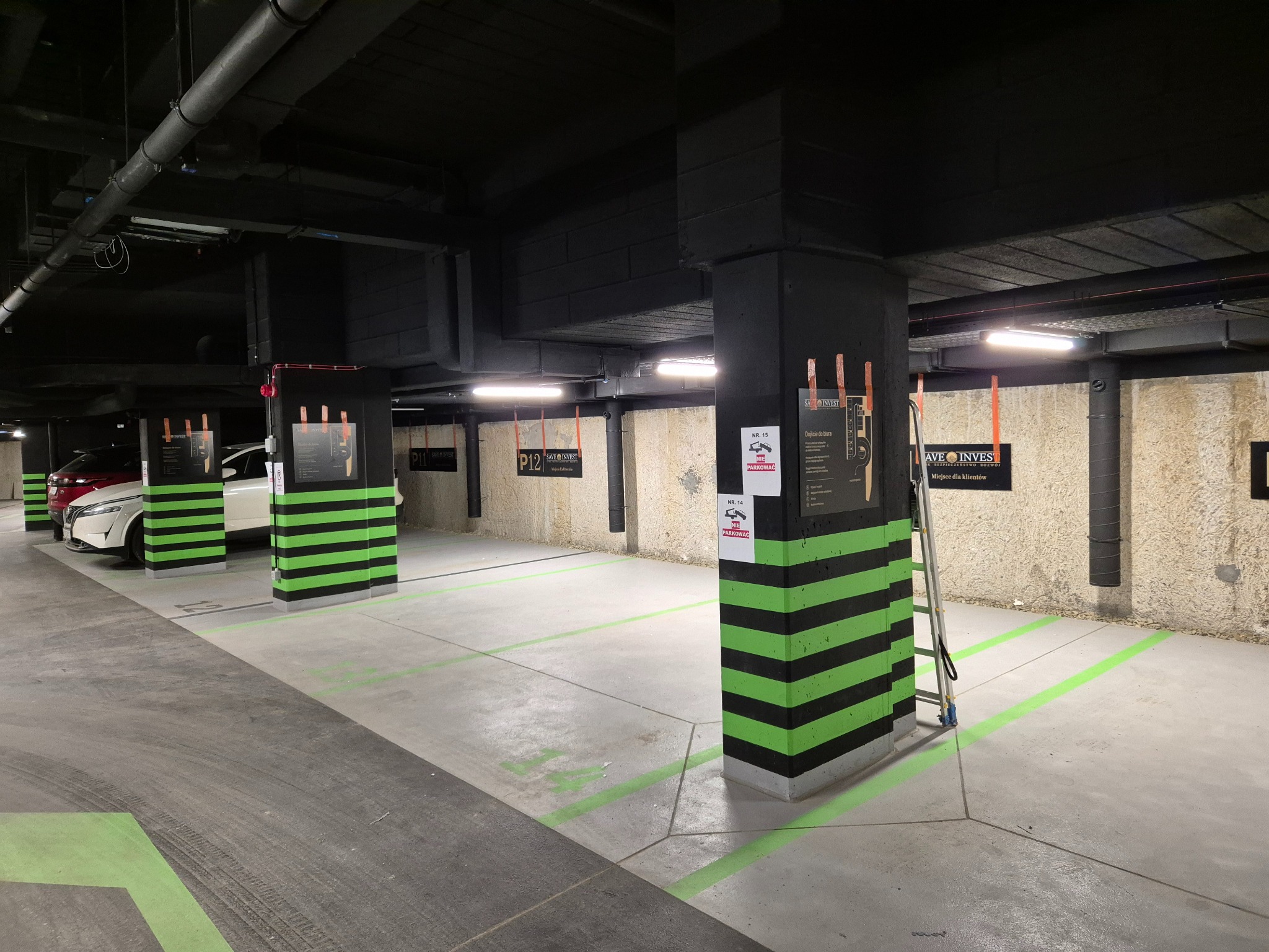 Oznakowane, podziemne miejsca parkingowe z kolumnami w czarno-zielone pasy, strzałki kierunkowe na posadzce, tabliczki informacyjne na kolumnach, widoczne rury pod sufitem.