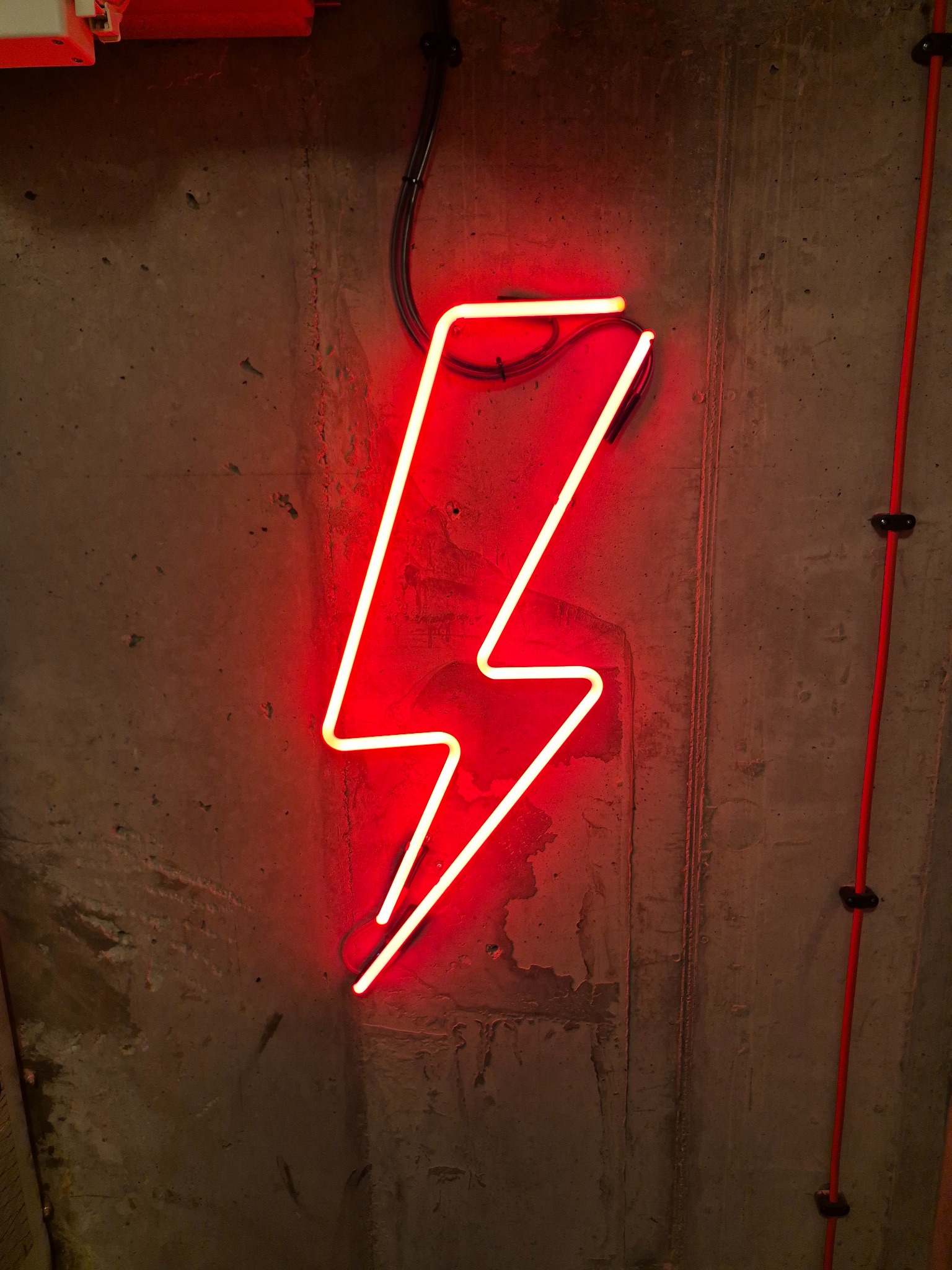 Czerwony neon w kształcie błyskawicy na surowej betonowej ścianie w Krakowie, obok pionowa czerwona rurka instalacyjna.
