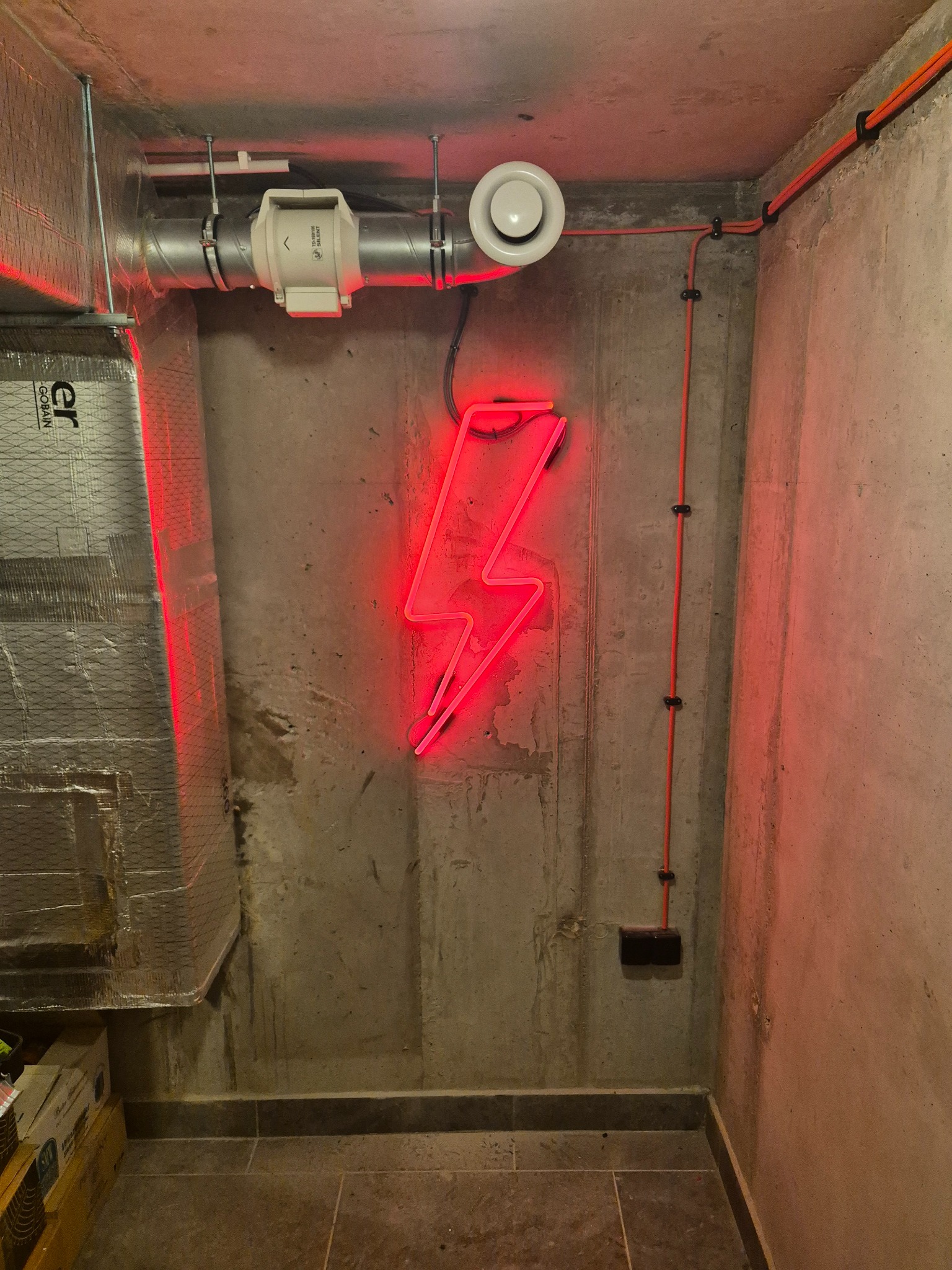 Betonowa ściana z neonowym znakiem błyskawicy, widoczne rury wentylacyjne, kable elektryczne w peszlach oraz fragment izolacji termicznej