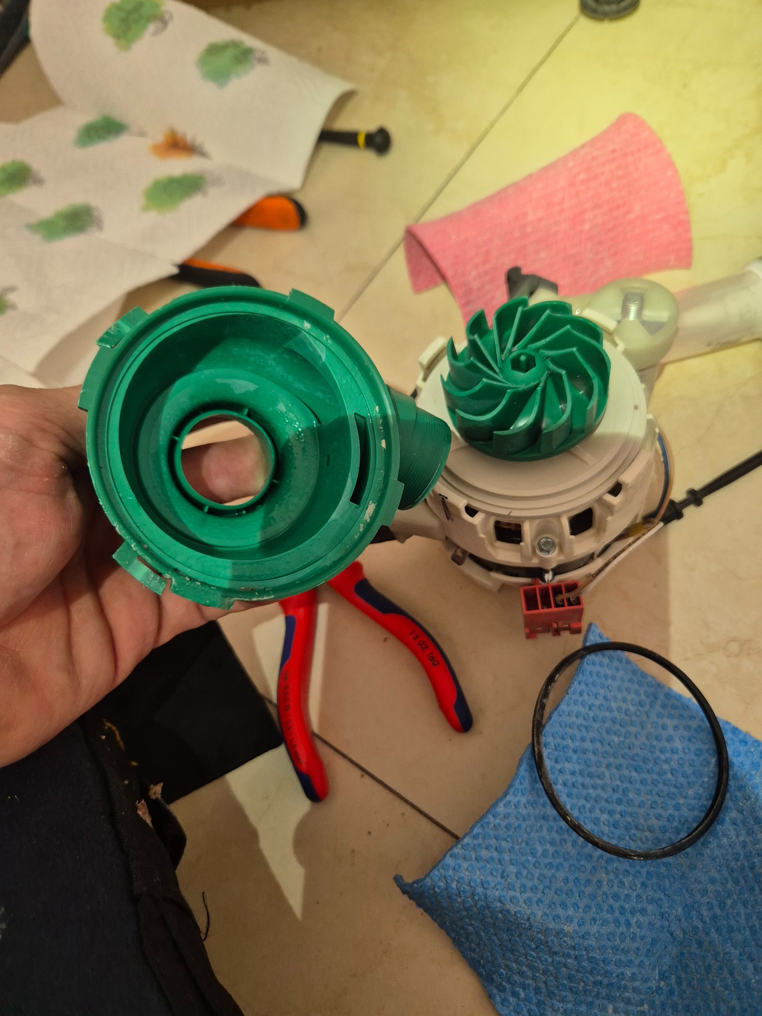 Rozłożona pompa zmywarki: zielona obudowa trzymana w dłoni, wirnik obok, widoczne narzędzia i uszczelka na niebieskiej ściereczce, przygotowane do montażu.