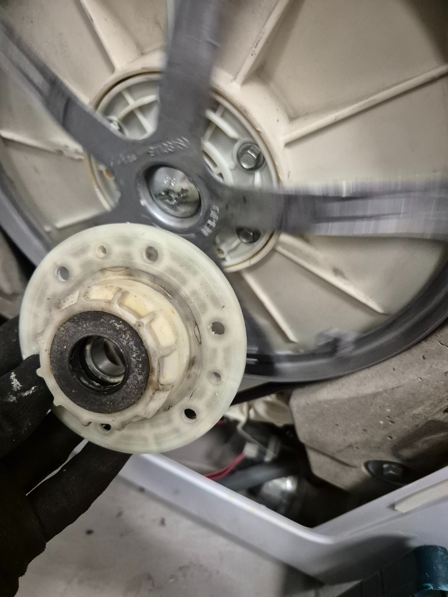 Naprawa pralki: dłoń w czarnej rękawicy trzyma plastikową tarczę z łożyskiem na tle bębna pralki podczas naprawy.