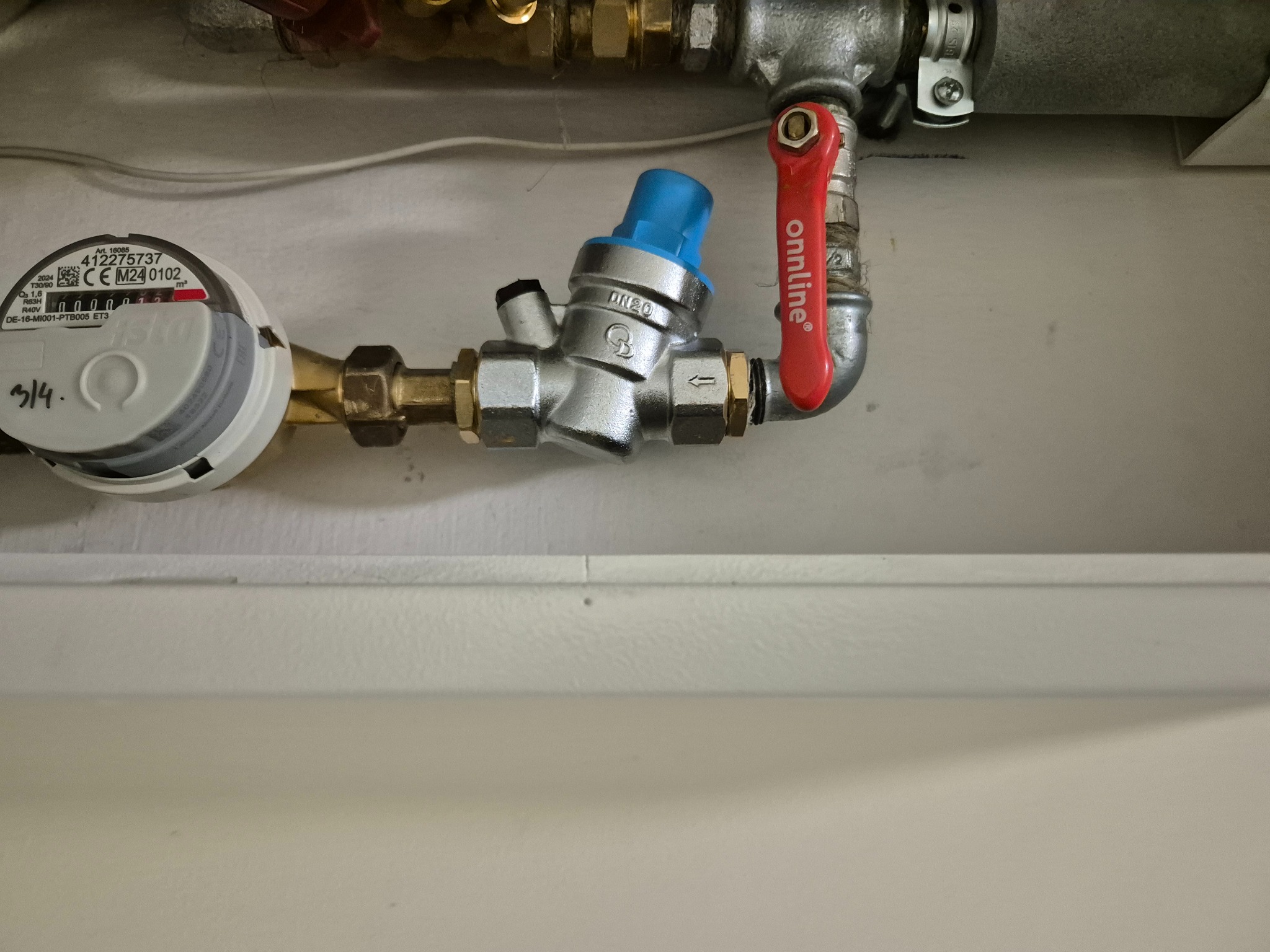 Zamknięty licznik wody z niebieskim regulatorem ciśnienia i czerwonym zaworem odcinającym marki Onnline, zamontowany na białej ścianie.